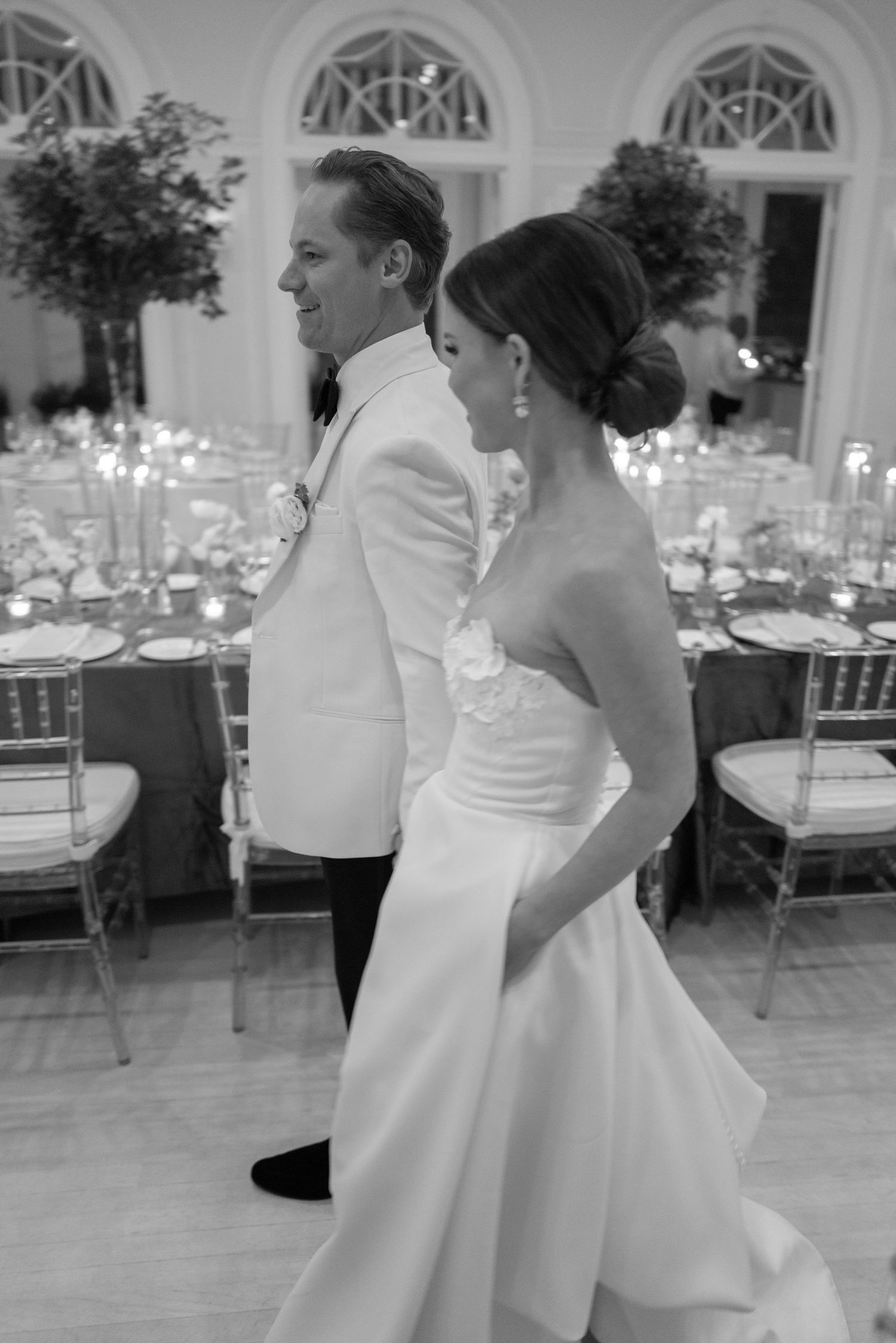 Black and white photo of bride in strapless gown and groom in white tuxedo at reception.
