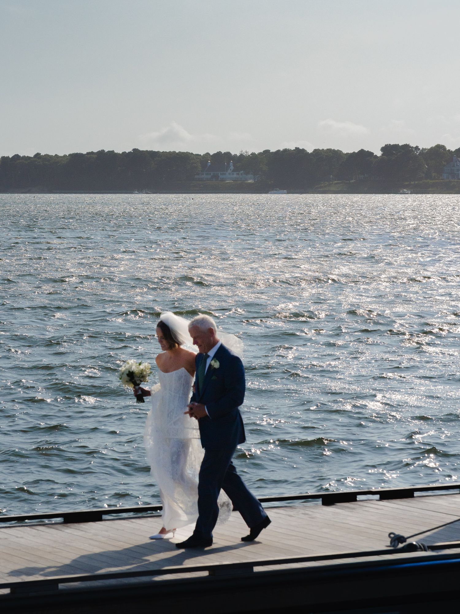 Bride and escort walking along dock with sparkling water in background at waterfront wedding.