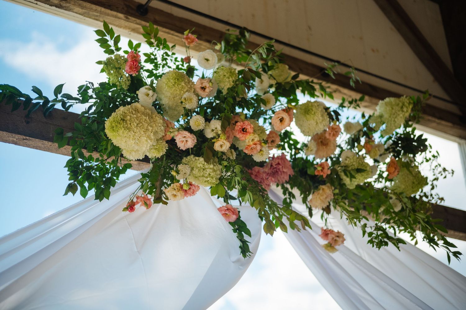 Floral wedding arch adorned with green hydrangeas, pink roses, and white draping against a blue sky.