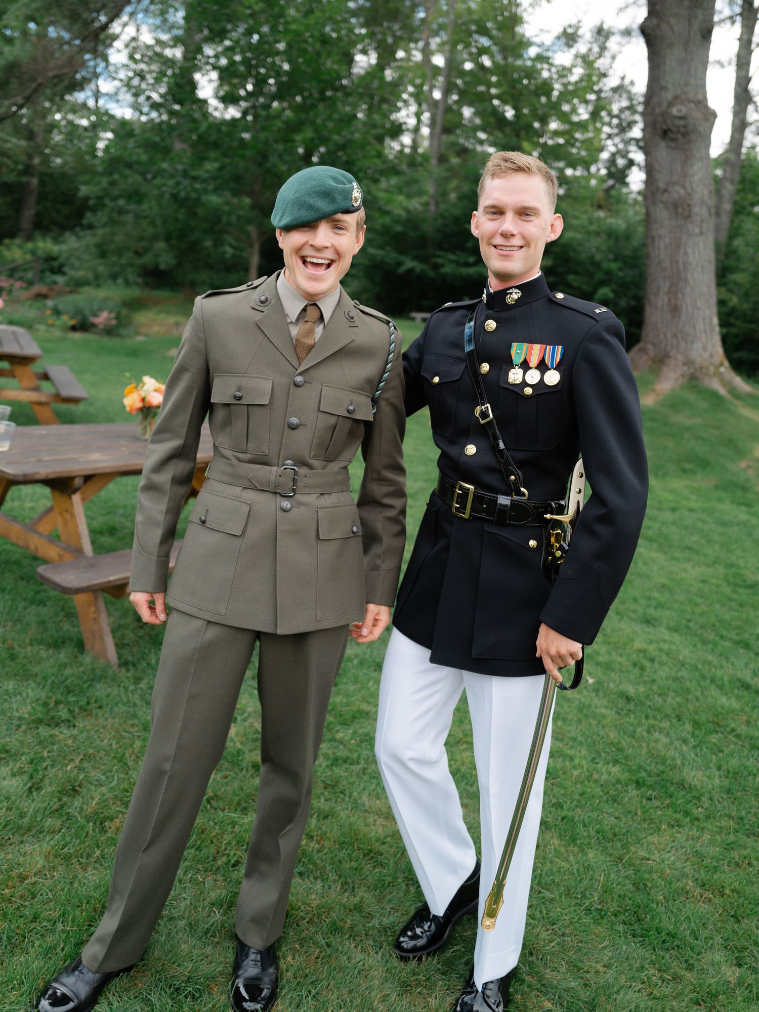 Two military personnel in dress uniforms smile outdoors at a wedding celebration.