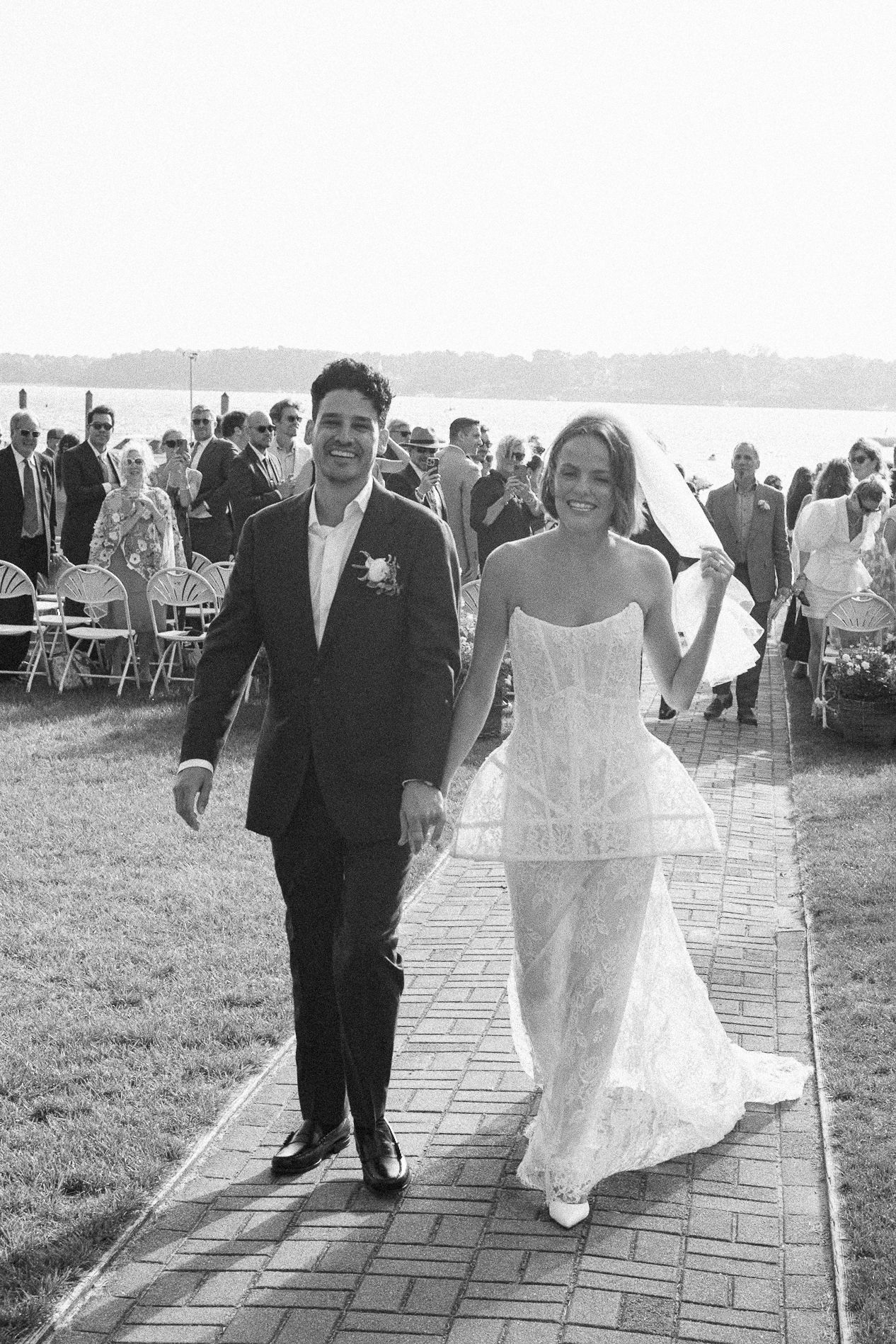 Black and white photo of a smiling bride and groom walking down the aisle after their outdoor waterfront wedding ceremony.