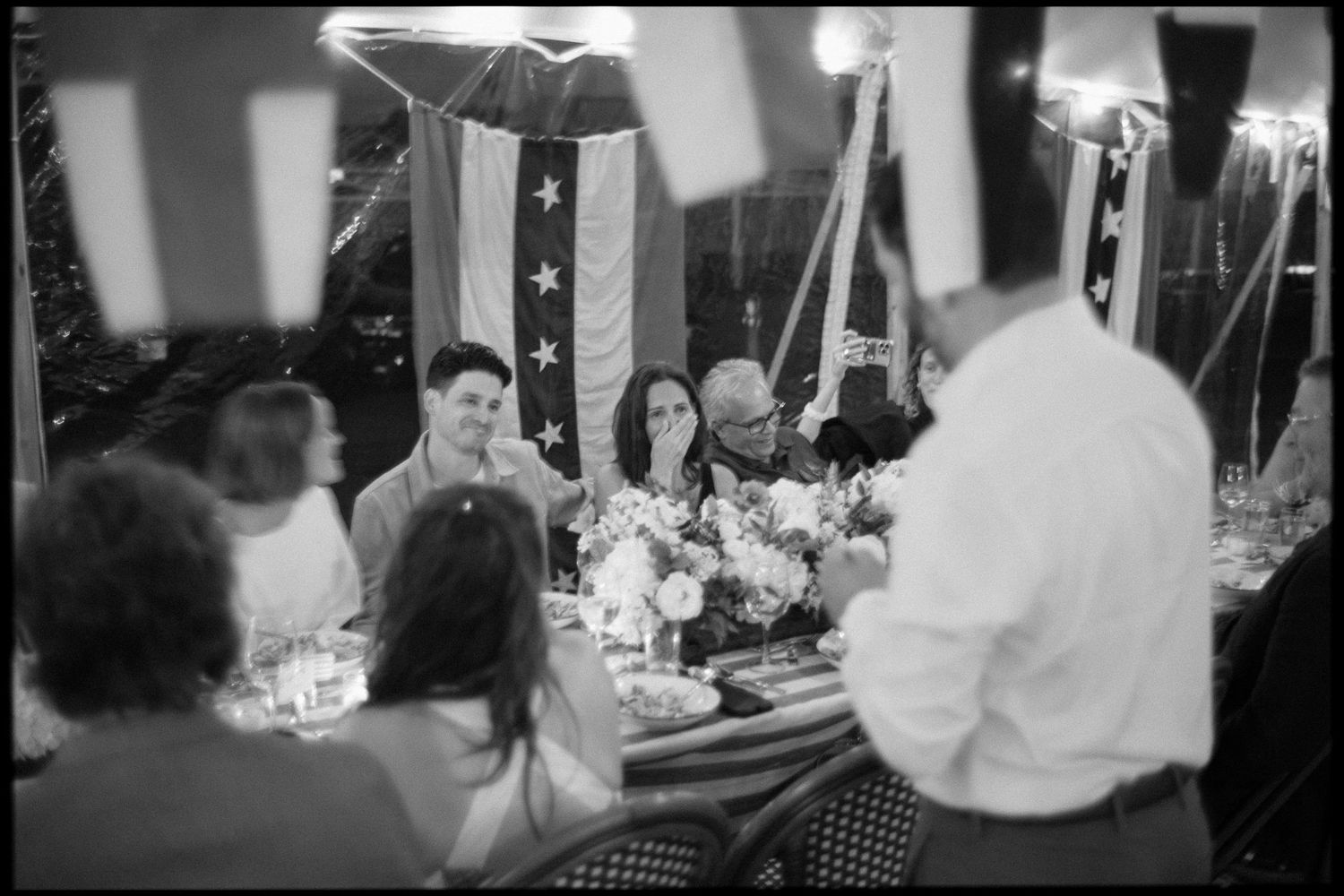 Black and white photo of wedding guests seated at floral-decorated dinner table under nautical flags.