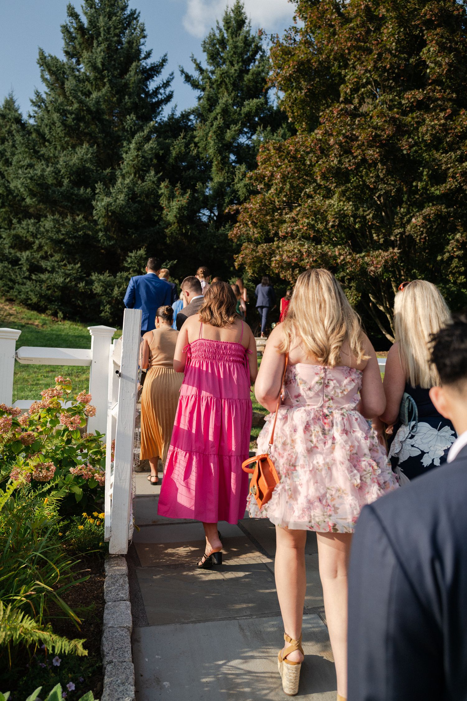 Wedding guests in colorful dresses walking along a garden path lined with flowers and trees.