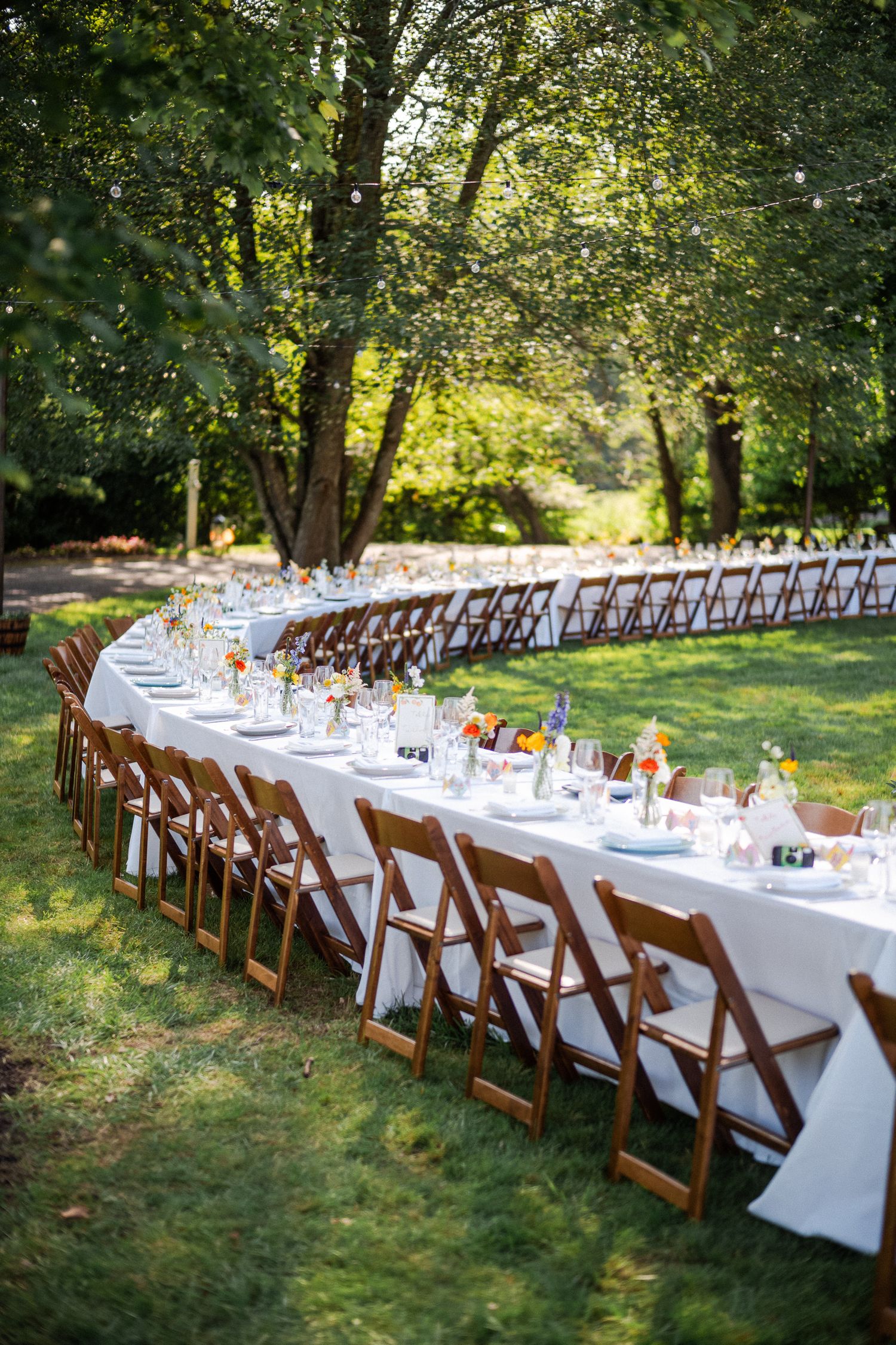 Long outdoor wedding reception tables with white linens and wooden folding chairs set under lush green trees.