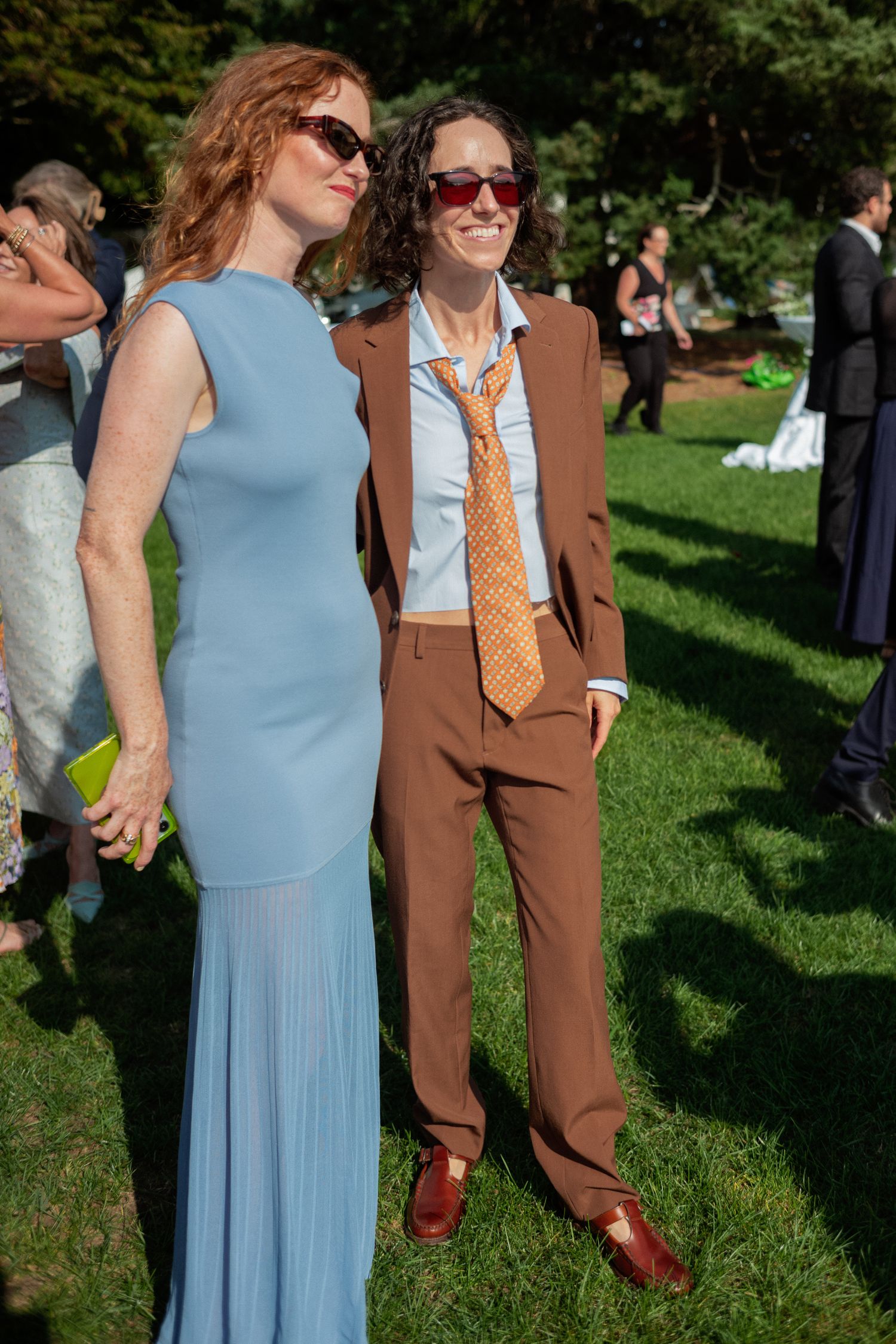 Two wedding guests pose on grass, one in blue gown, other in brown suit with orange tie and sunglasses.