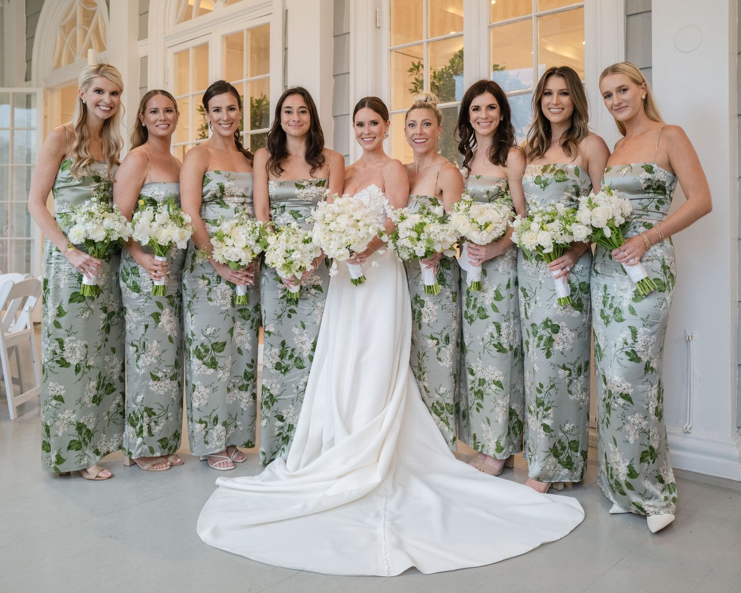 Bride in white gown with bridesmaids in floral green dresses holding white bouquets at elegant venue.