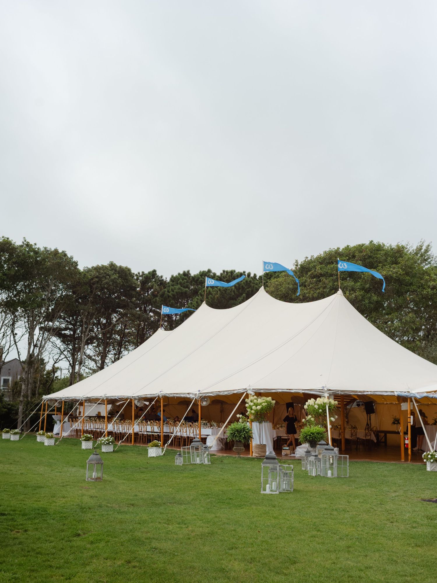 Large white sailcloth wedding tent with blue flags on green lawn under overcast sky.