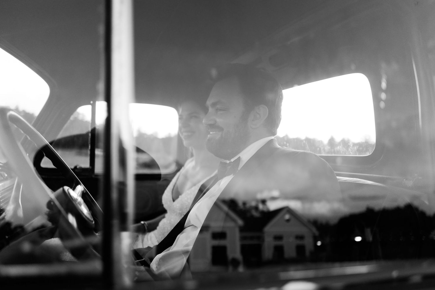 Smiling groom in formal attire drives a vintage vehicle at a wedding, photographed through the windshield.