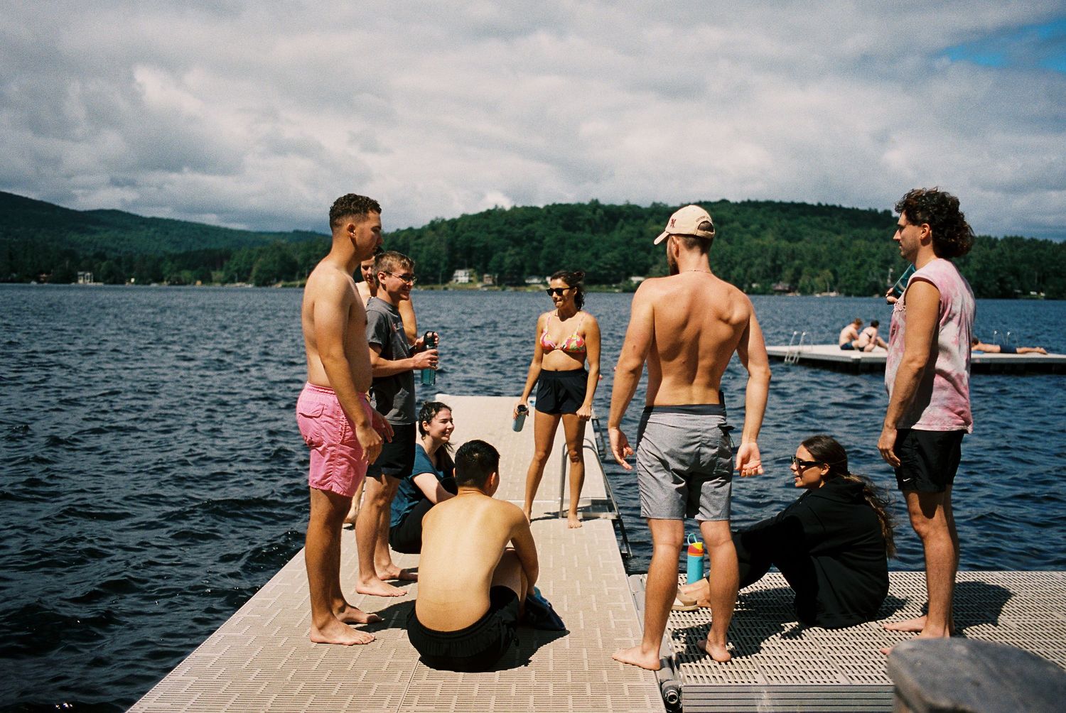 Group of young adults in swimwear socializing on a lake dock with mountains in background.
