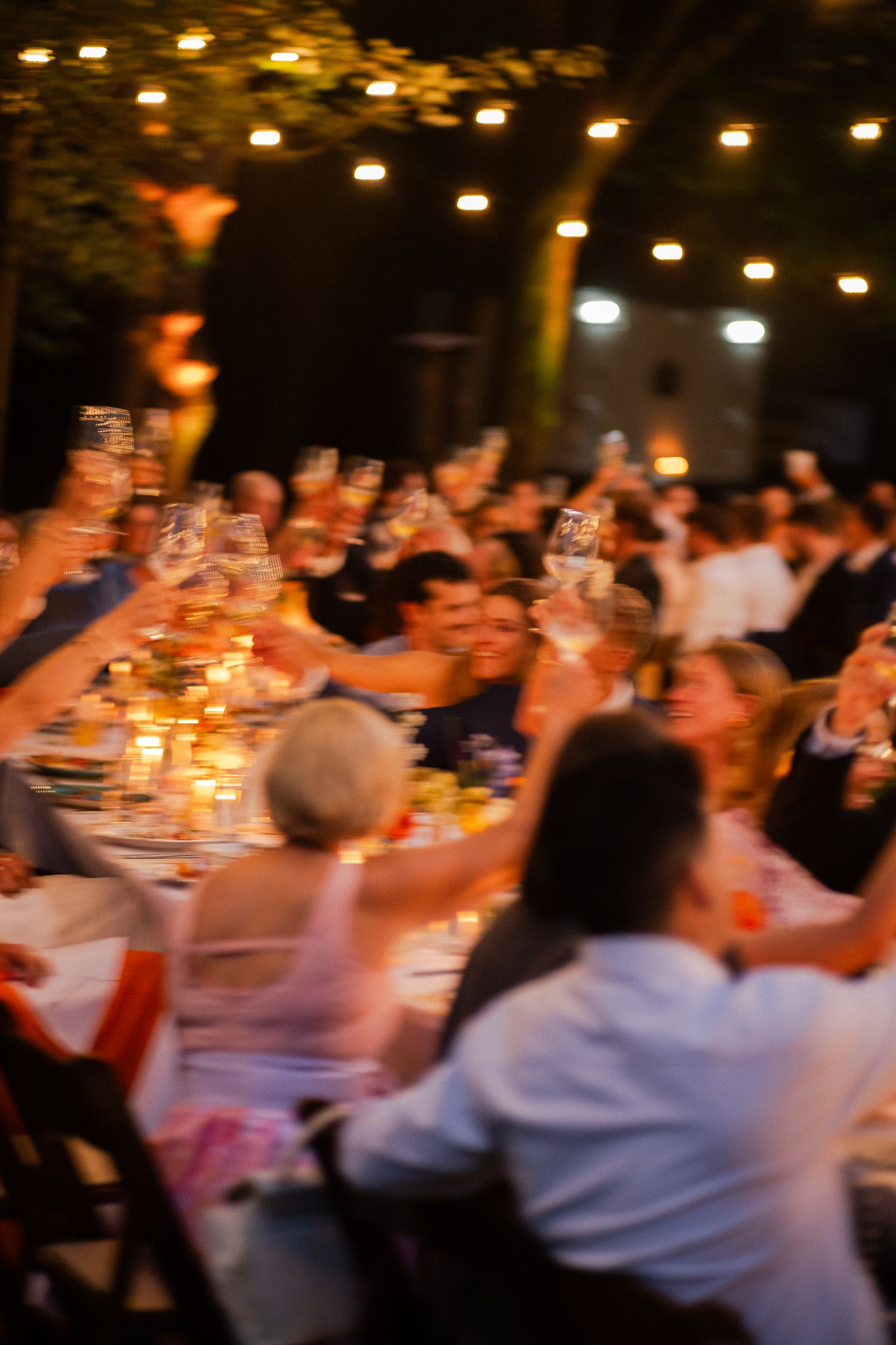 Blurred photo of wedding guests raising glasses in a joyful outdoor evening toast.