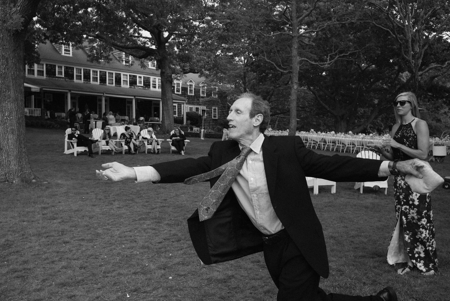 Man in suit strikes dramatic pose on lawn at outdoor wedding reception in black and white photo.