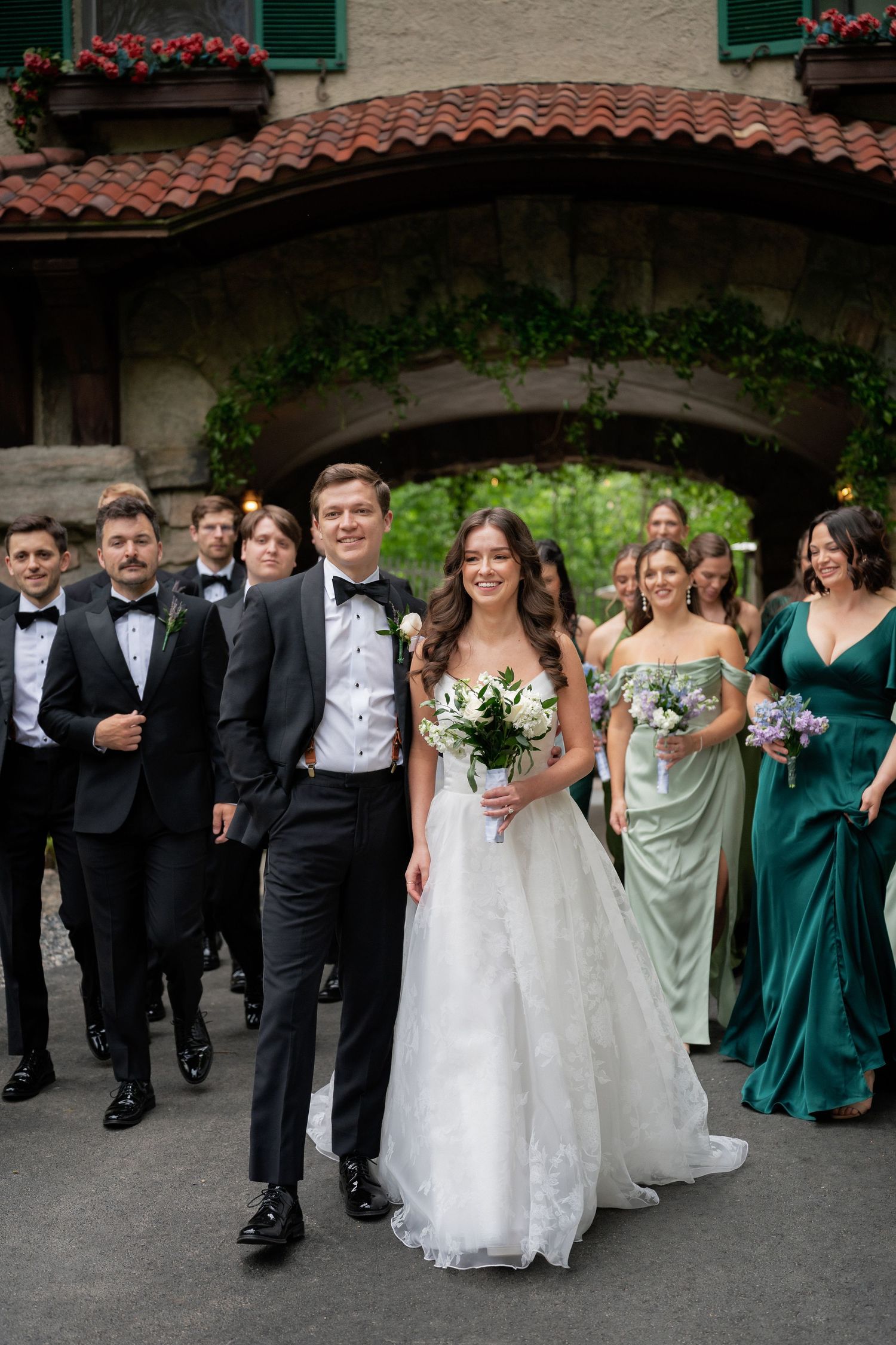 Wedding party walks together under stone arch with bridesmaids in sage green and emerald dresses.