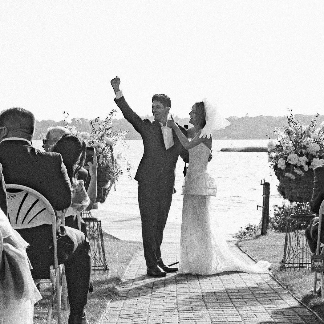 Black and white photo of a joyful couple celebrating at an outdoor waterfront wedding ceremony with guests seated nearby.