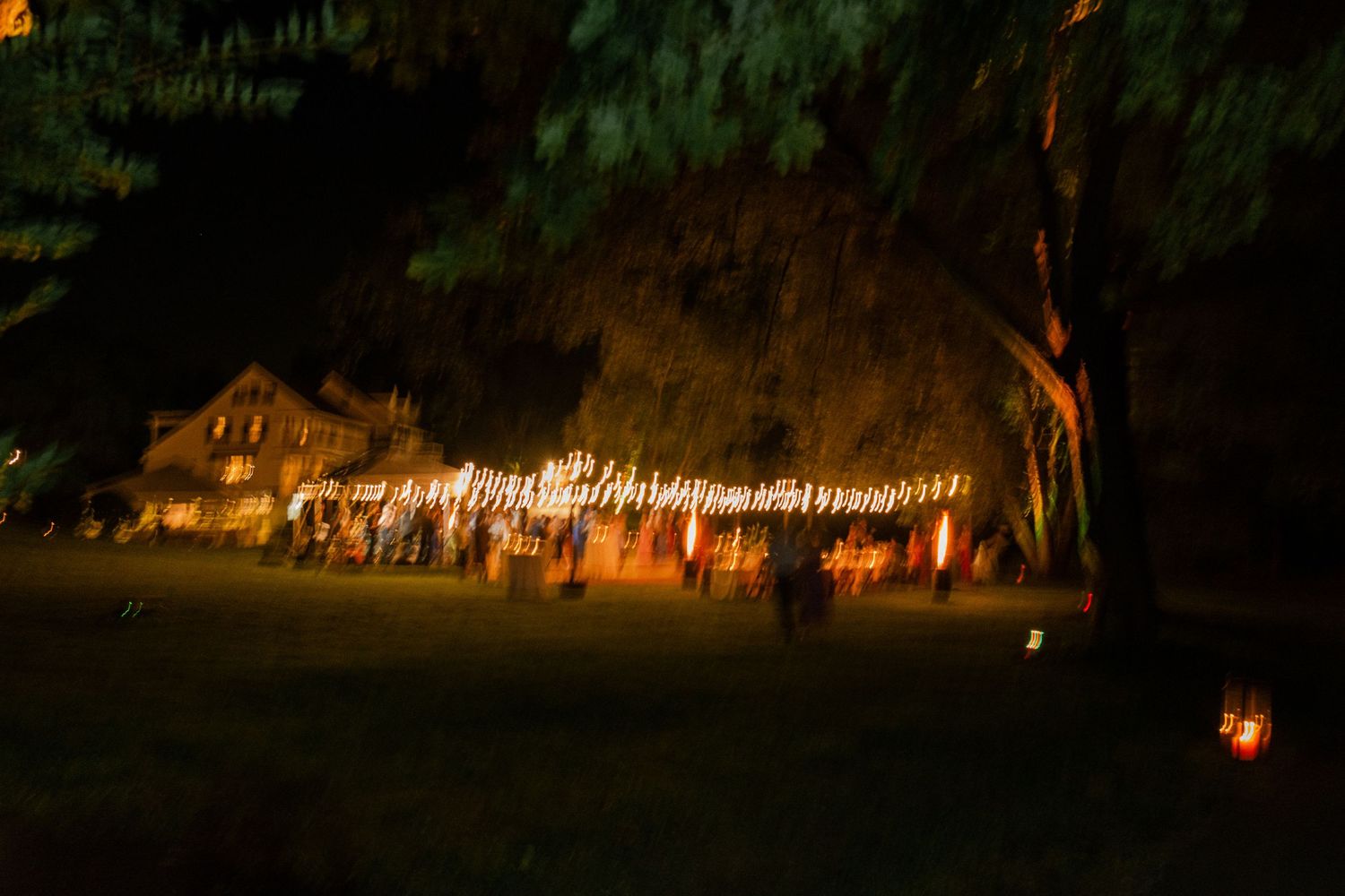 Colorful blurred motion shot of outdoor wedding reception with string lights glowing under large trees at night.