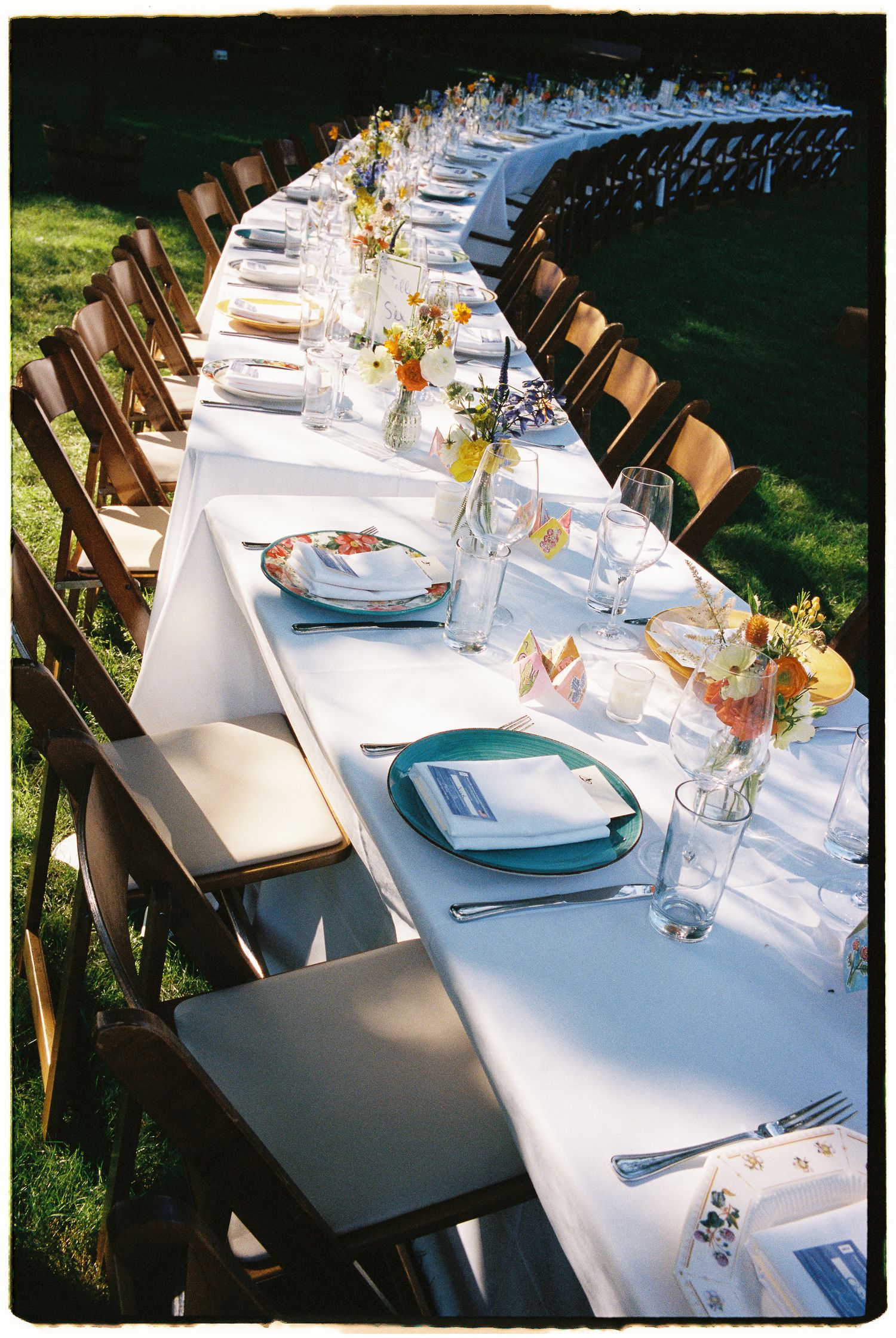 Long outdoor banquet table with white linen, colorful floral centerpieces, teal plates, and wooden folding chairs on green grass.