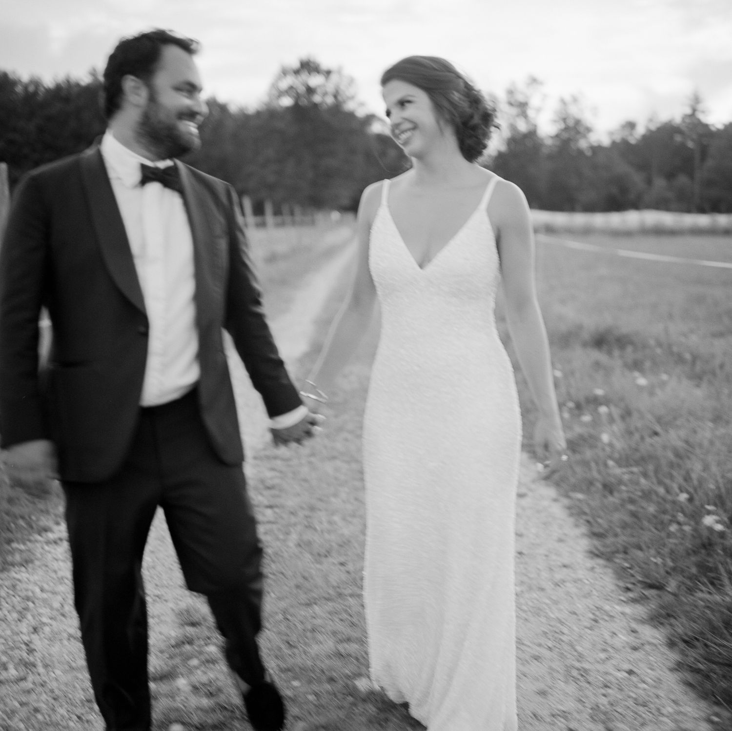 Black and white photo of bride and groom holding hands walking along scenic rural path at wedding.