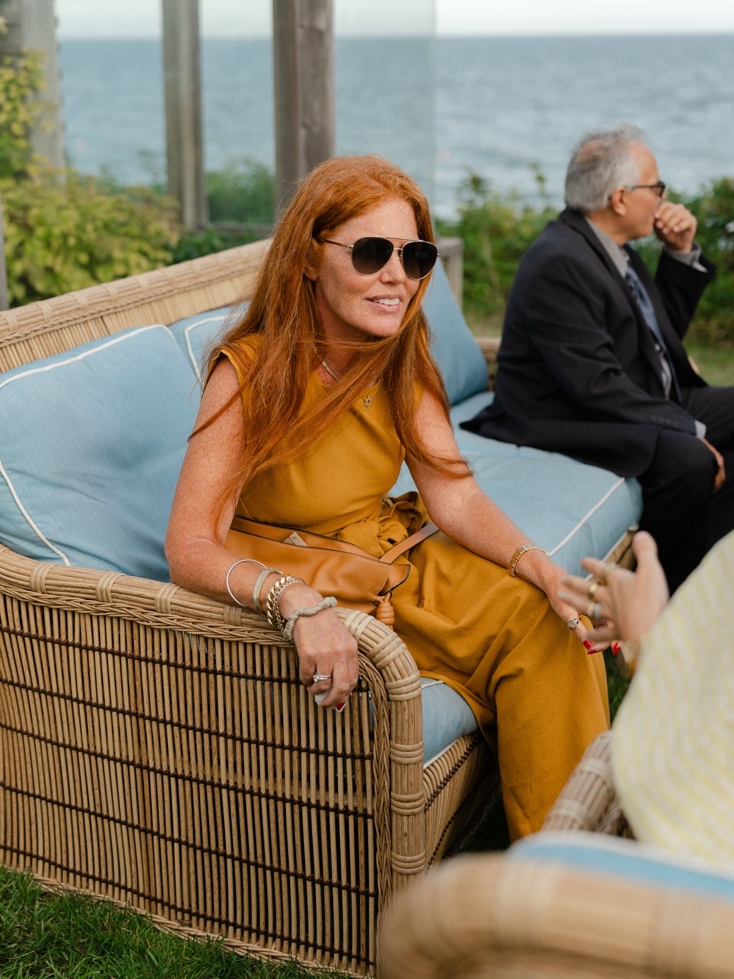 Stylish woman in mustard jumpsuit and sunglasses seated outdoors at waterfront wedding.