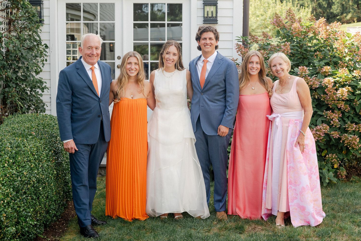 Wedding family portrait with bride in white gown and guests in coral and blue attire posing outdoors.