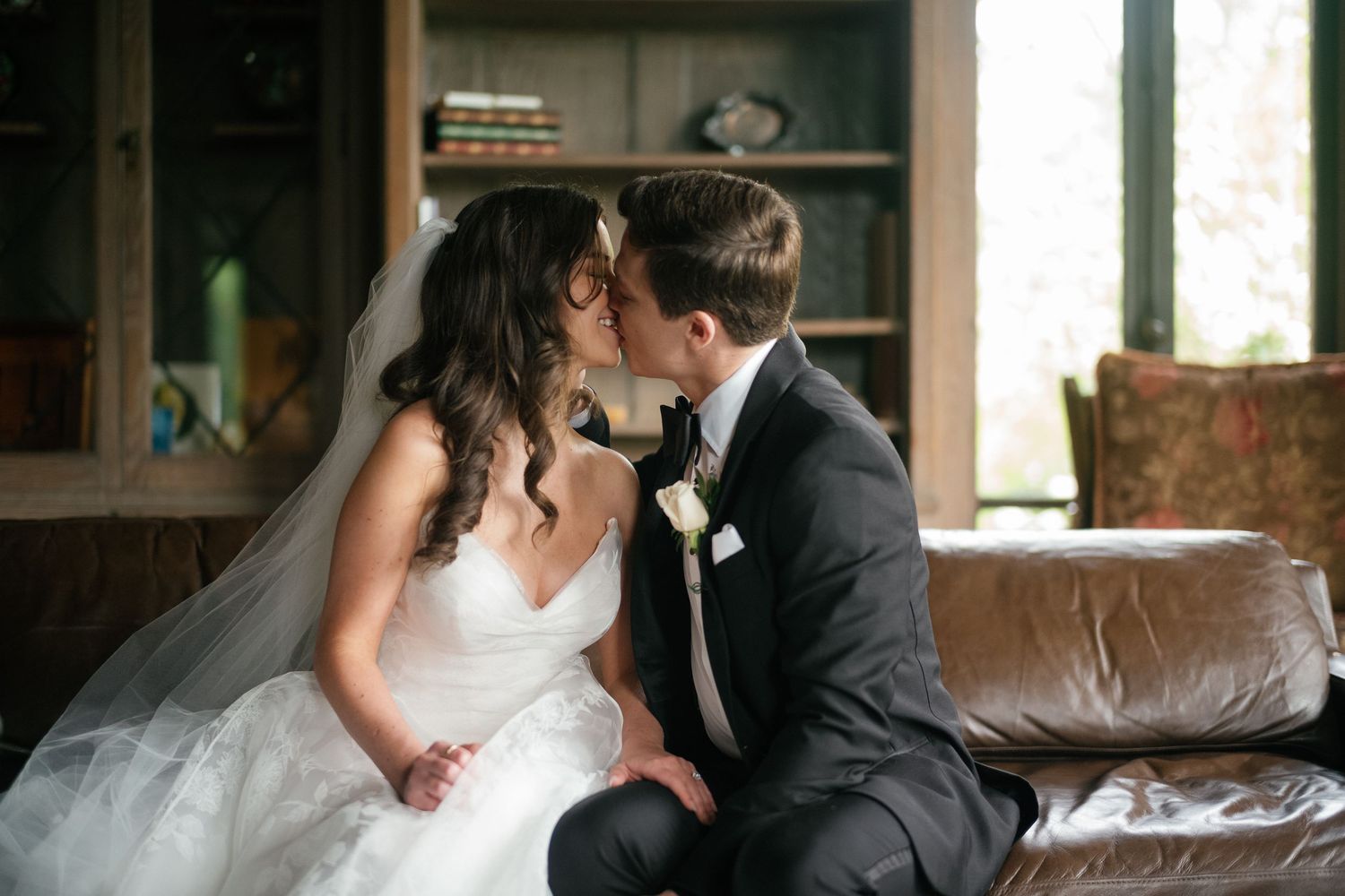 Bride and groom sharing a romantic kiss on a leather sofa during their wedding day indoor portrait session.
