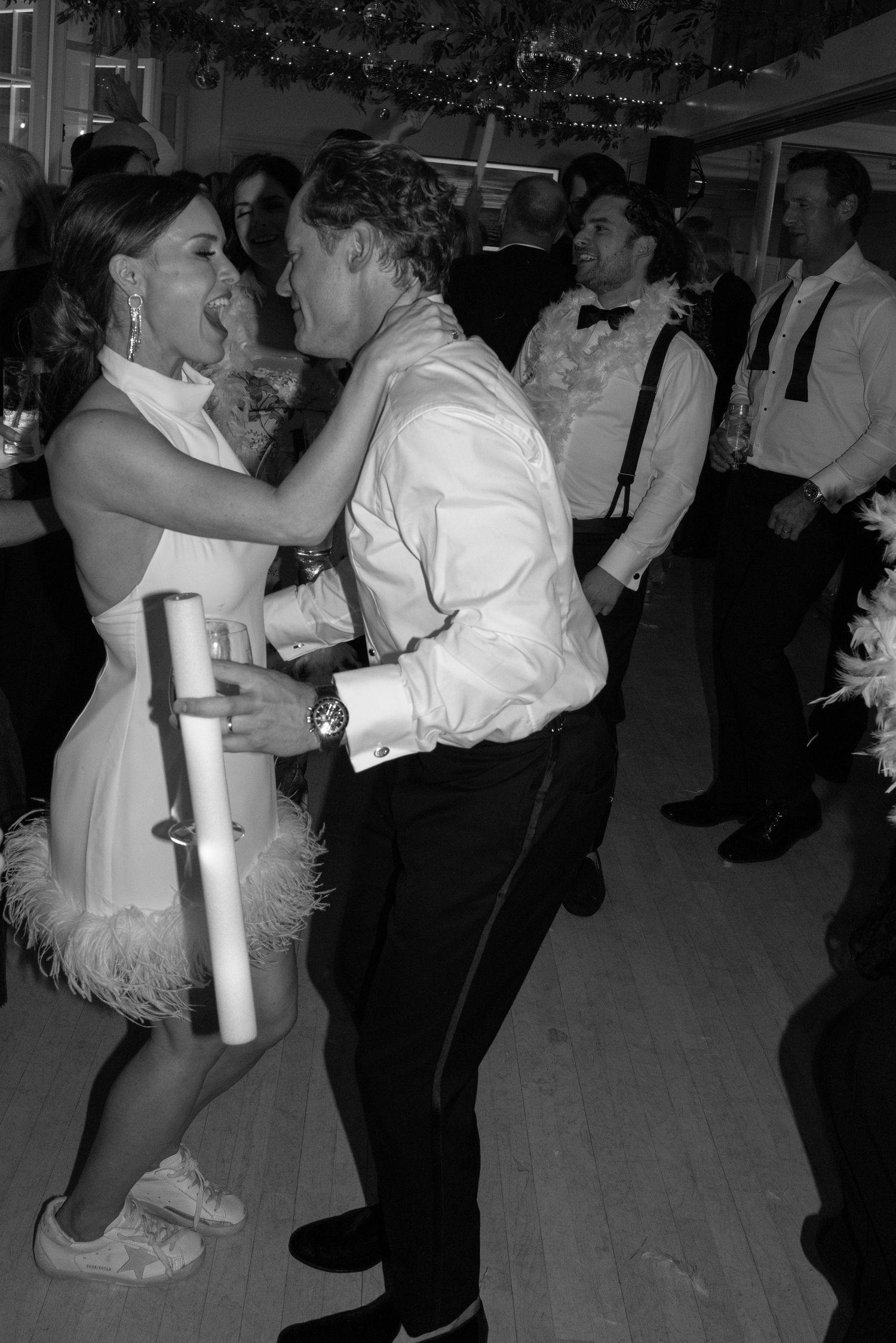 Black and white photo of bride in feathered dress dancing energetically with guest at wedding reception.