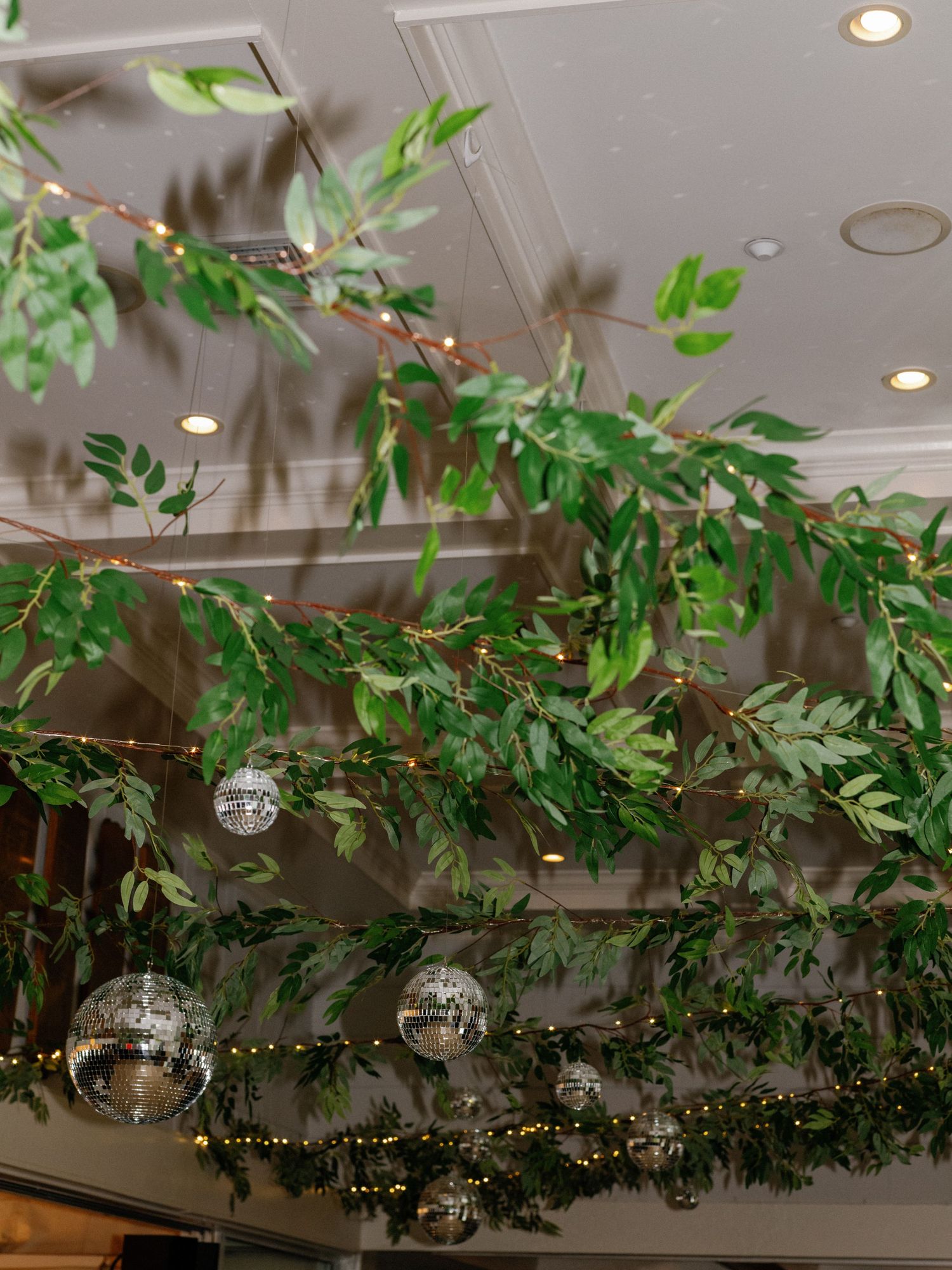 Wedding venue ceiling decorated with lush greenery garlands, fairy lights, and disco ball ornaments.
