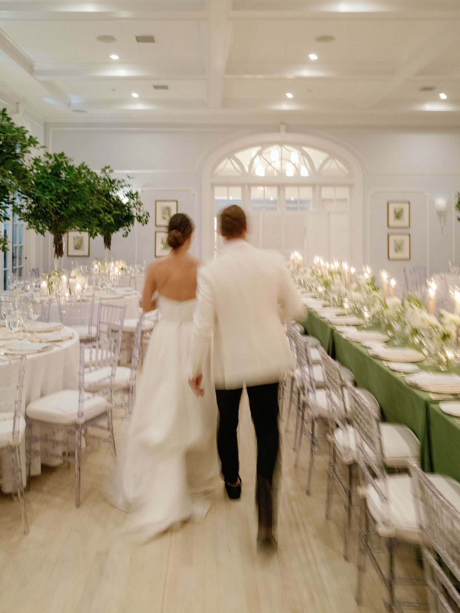 Bride and groom walking through elegantly decorated reception hall with tall greenery and candlelit tables.