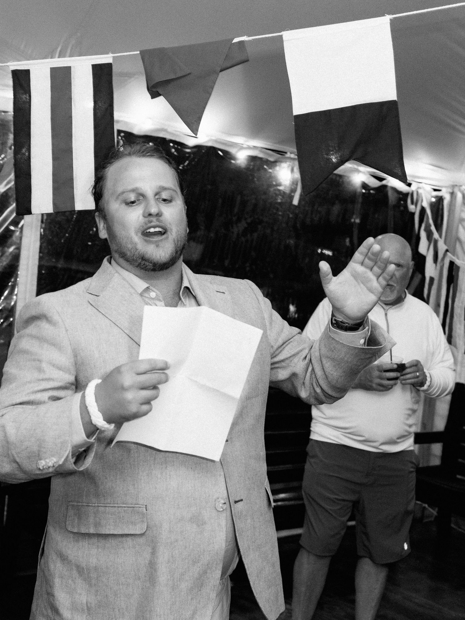 Man in blazer giving a toast speech holding paper under nautical flags at an outdoor wedding event.