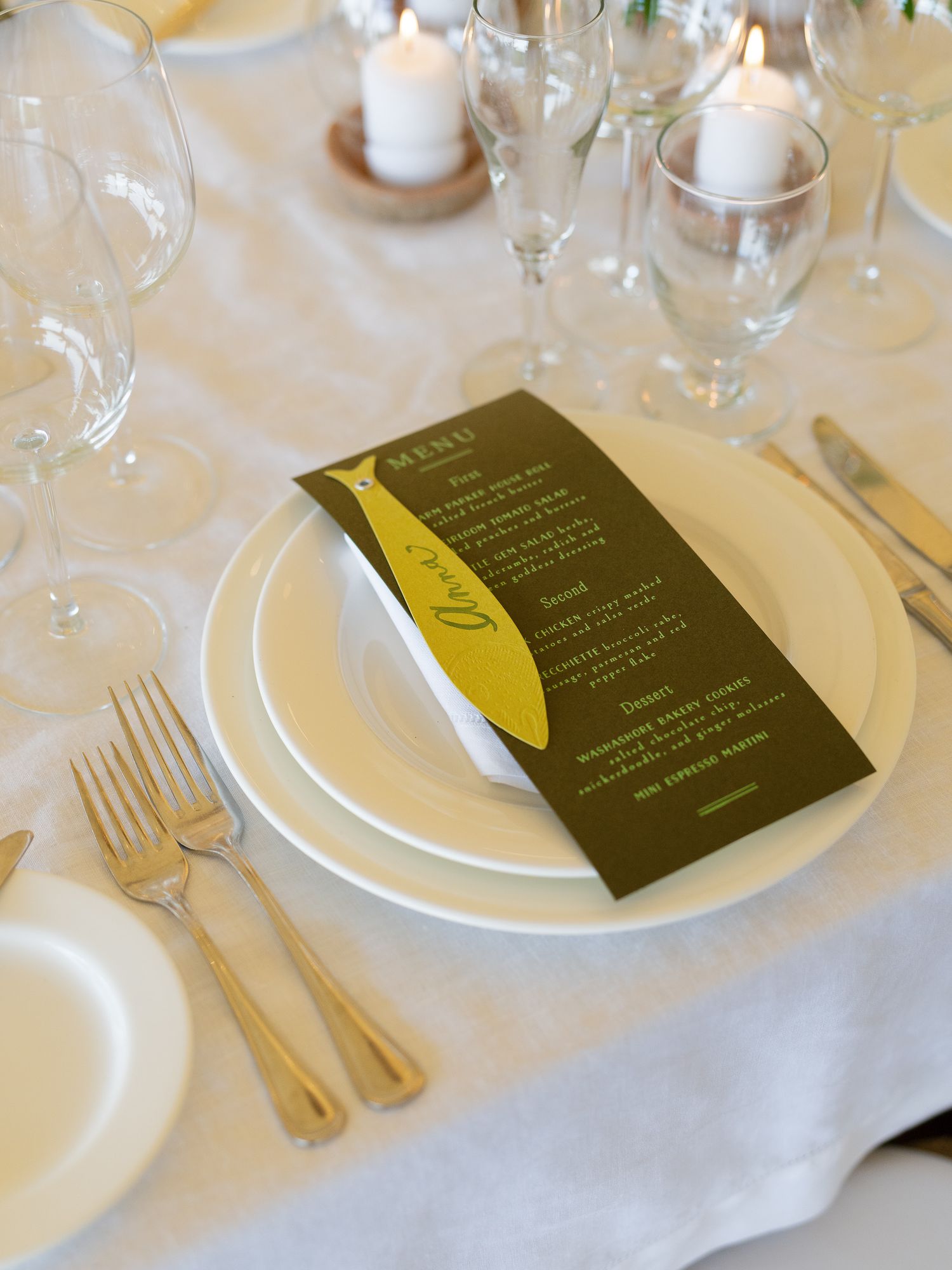 Wedding place setting with olive green menu card, white plates, and gold cutlery.