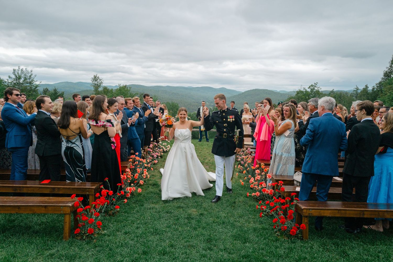 Newlyweds walk down outdoor aisle lined with red flowers as guests applaud with mountain scenery behind.