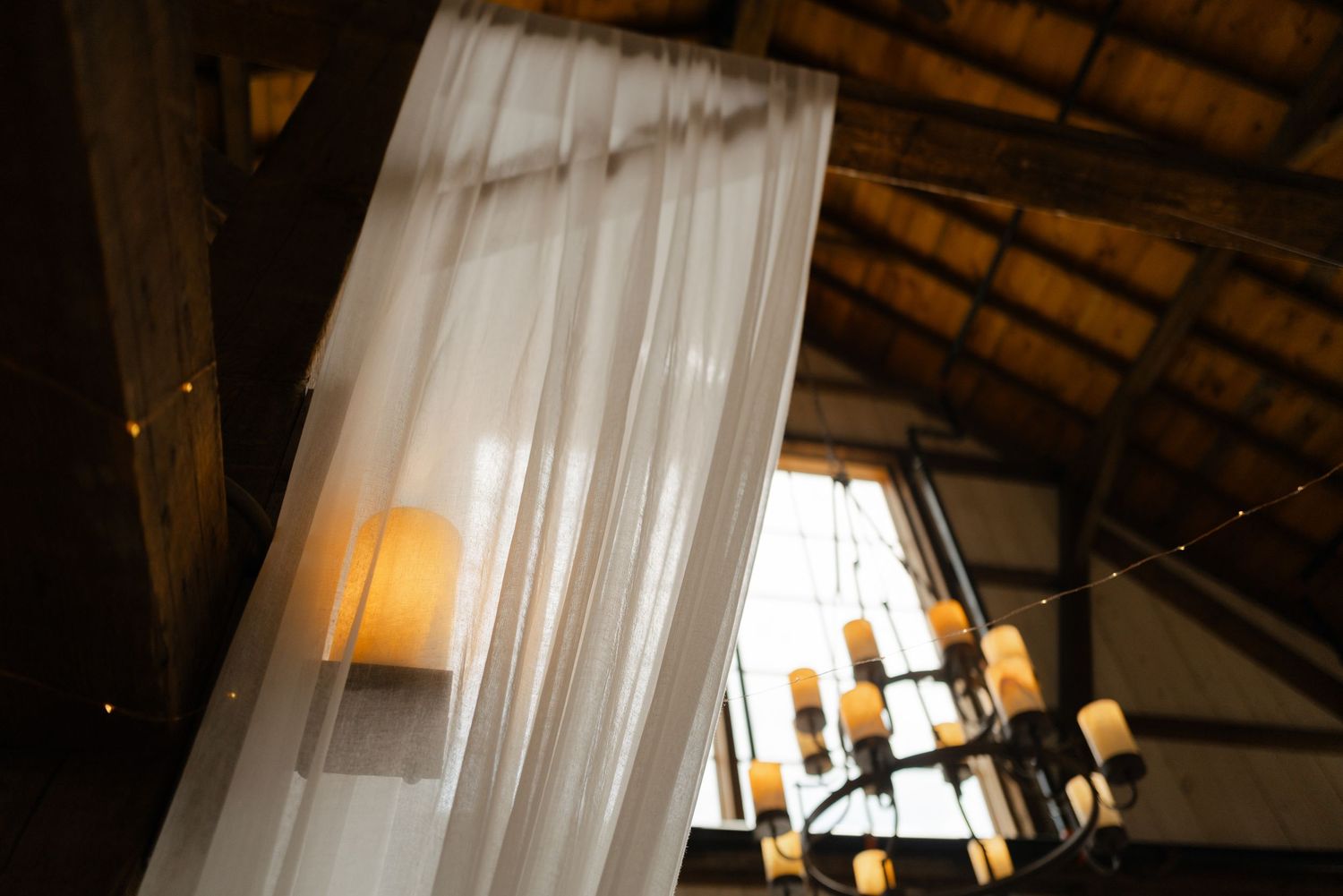 White sheer draping and rustic chandeliers in barn wedding venue ceiling.