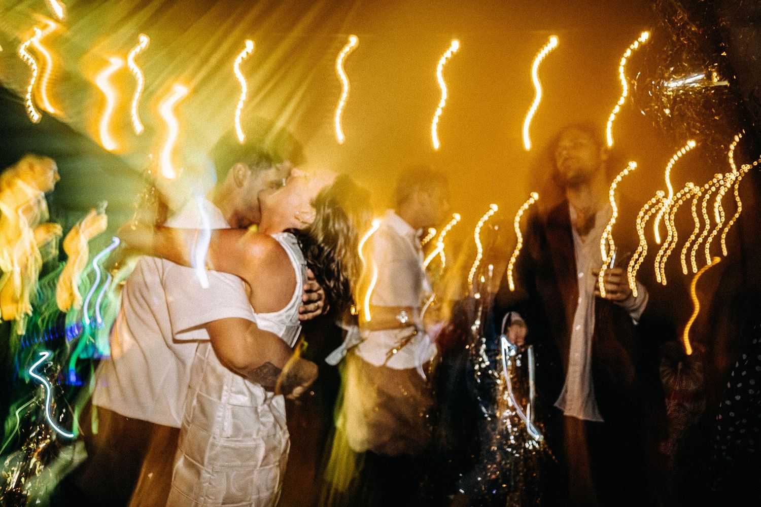 Couple embraces on dance floor with bokeh light streaks in gold and white at a lively wedding reception.