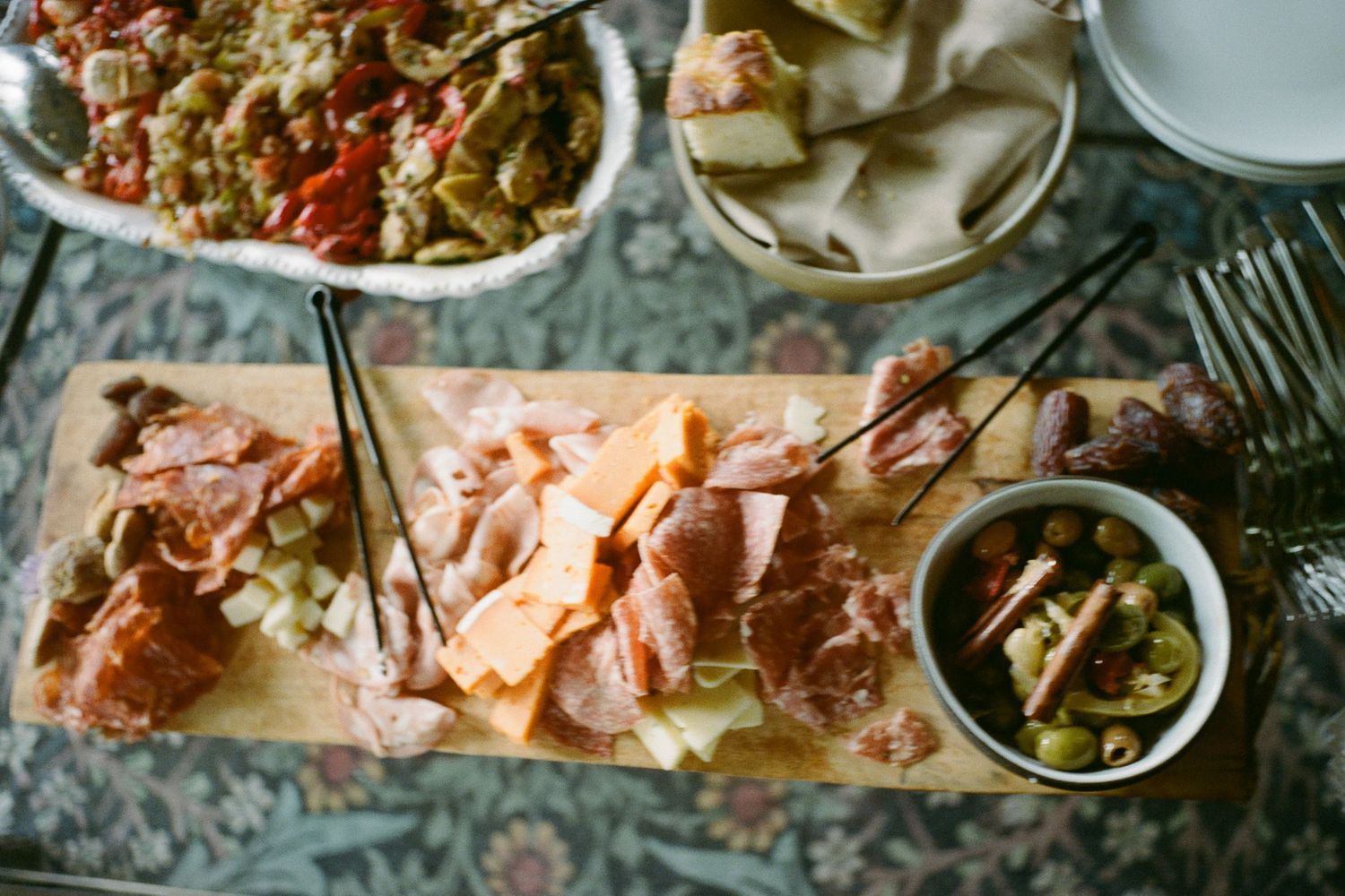 Charcuterie board with cured meats, cheese, and olives alongside salads on a floral tablecloth.