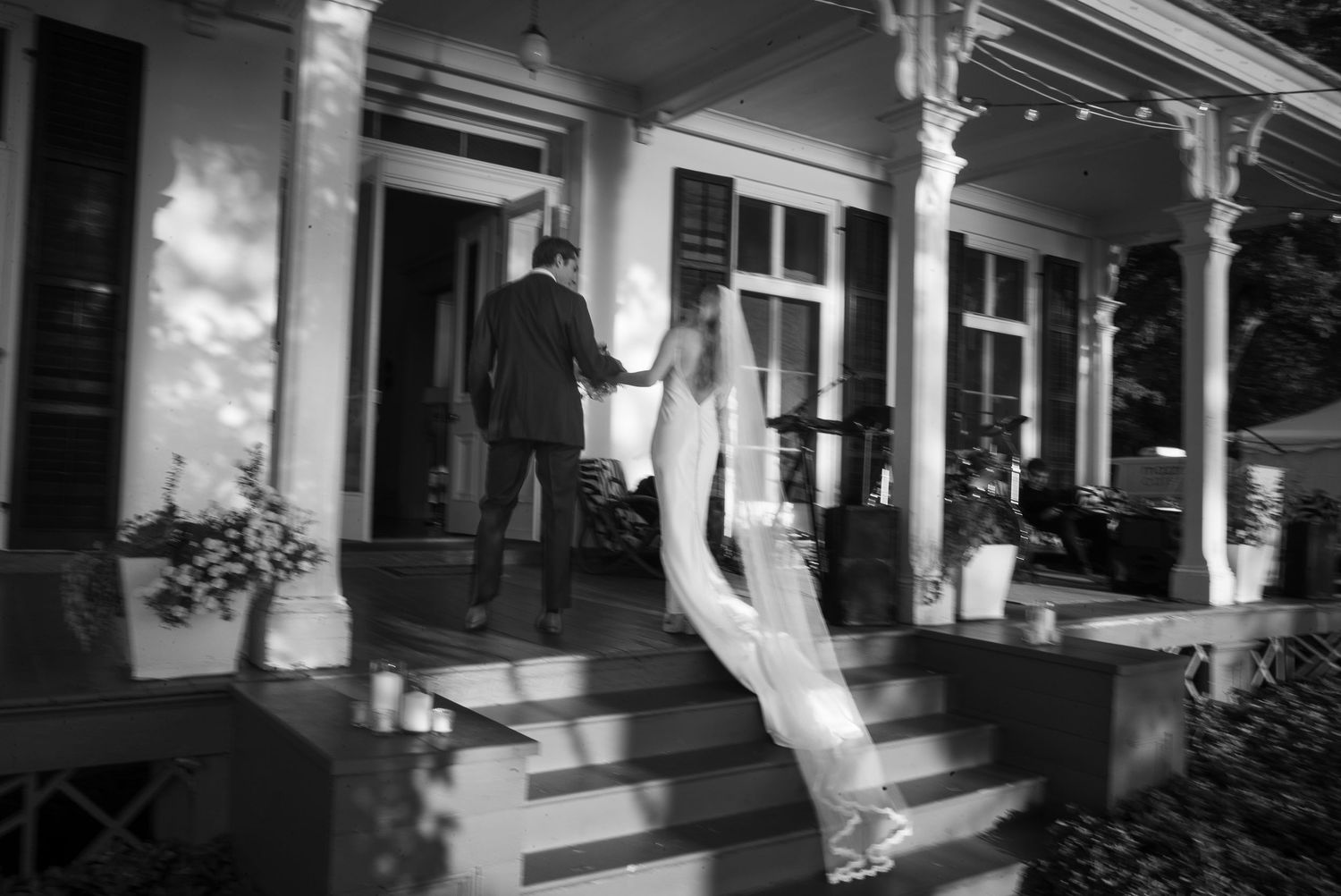 Black and white photo of bride and groom holding hands walking up steps to elegant venue porch.