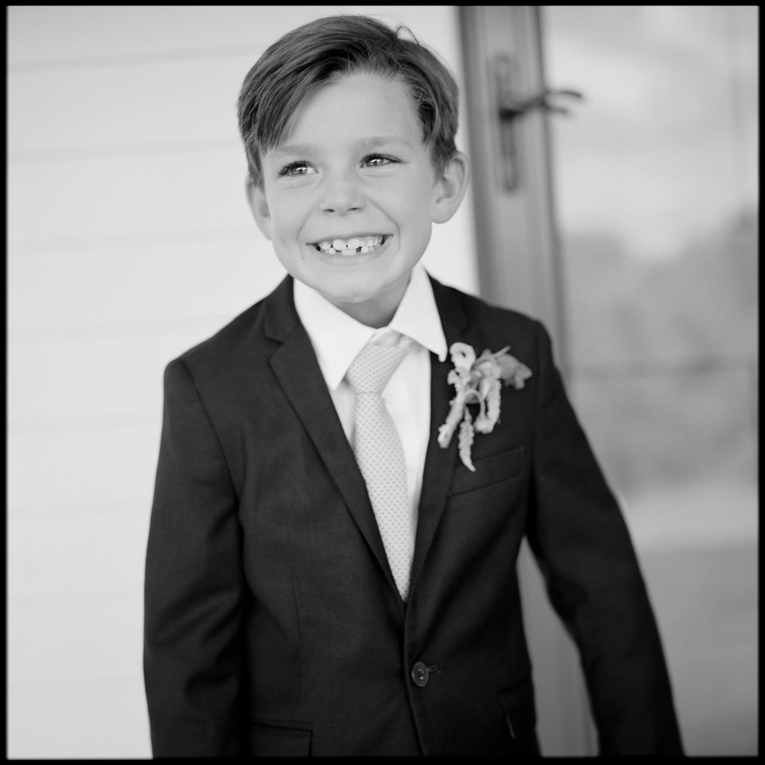 Adorable young boy in dark suit and boutonniere smiling at a wedding in black and white photo.