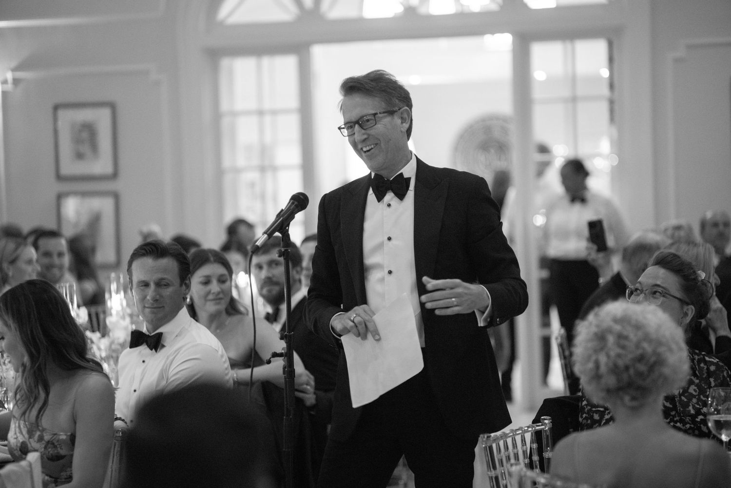 Elegant man in tuxedo delivering a speech at a formal wedding reception in a grand ballroom.