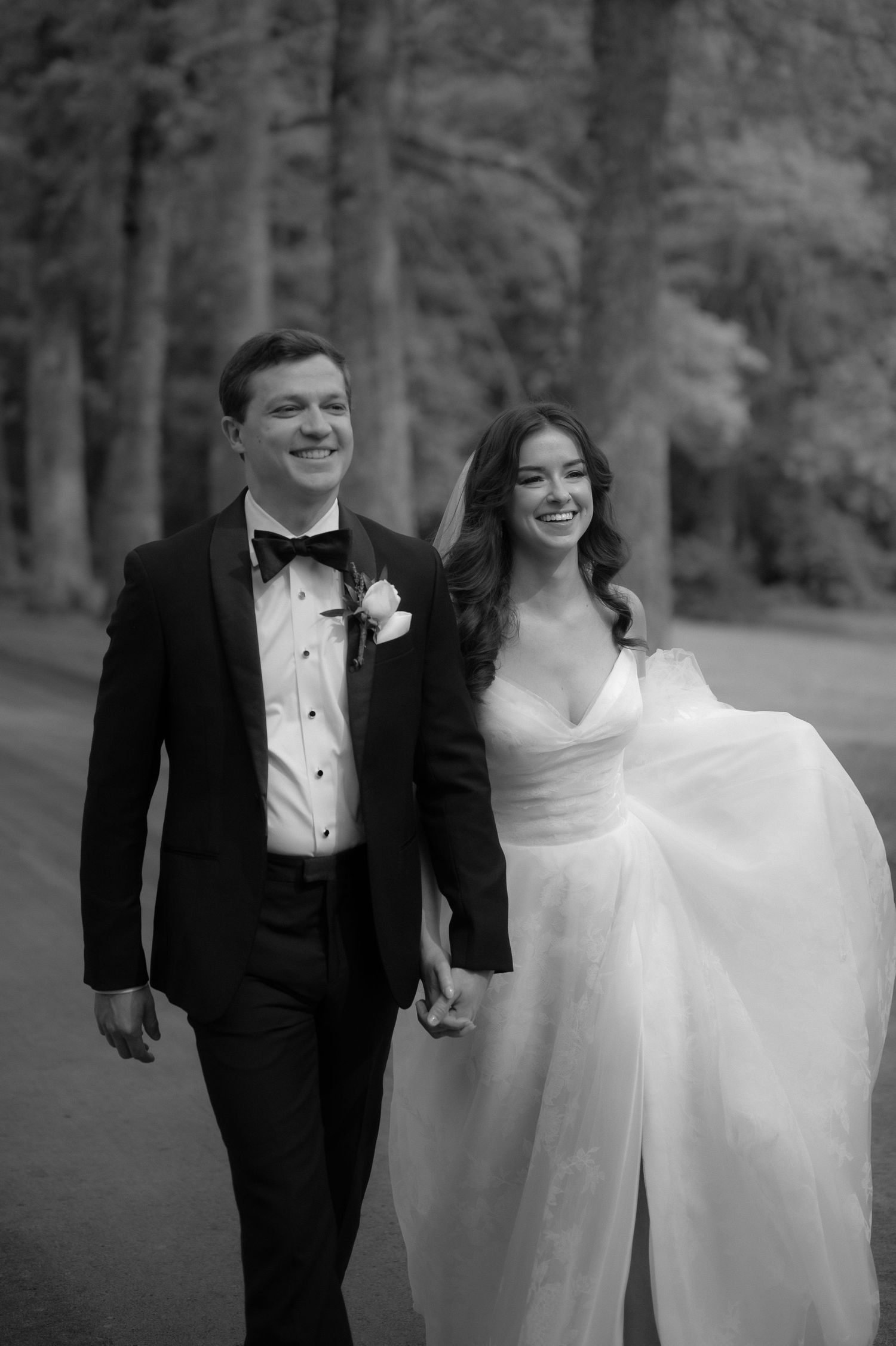 Black and white photo of smiling bride and groom walking hand in hand through trees.