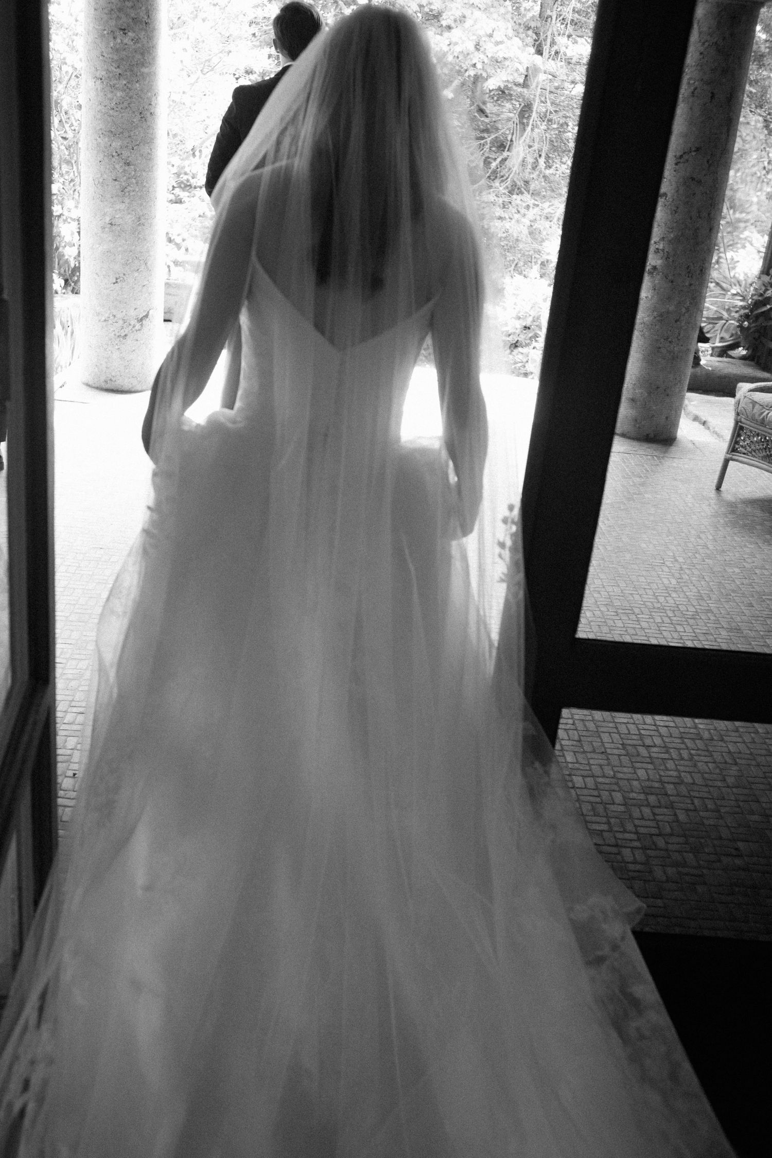 Black and white rear view of a bride in a white gown and veil walking toward light.