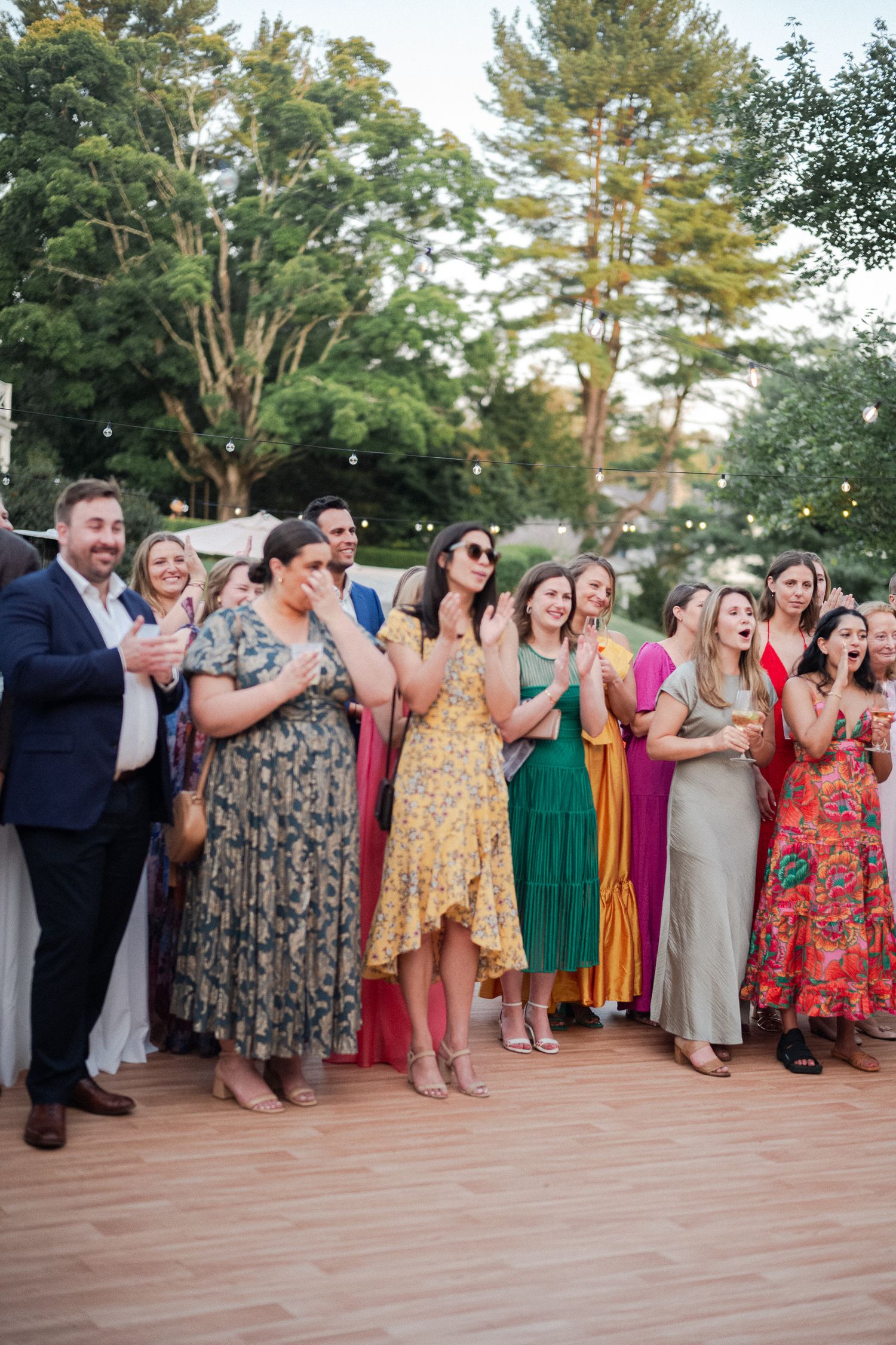 Wedding guests in colorful dresses react with joy and applause during outdoor reception celebration.