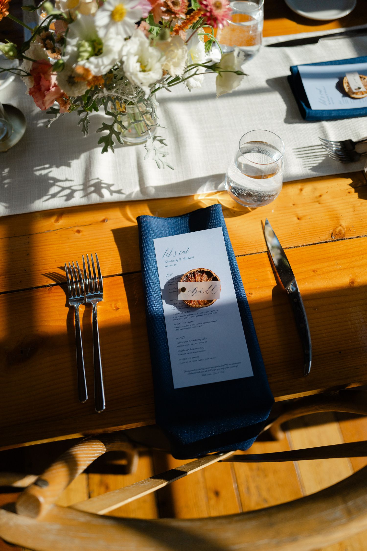 Elegant wedding place setting with blue napkin, printed menu, silverware, and floral centerpiece.