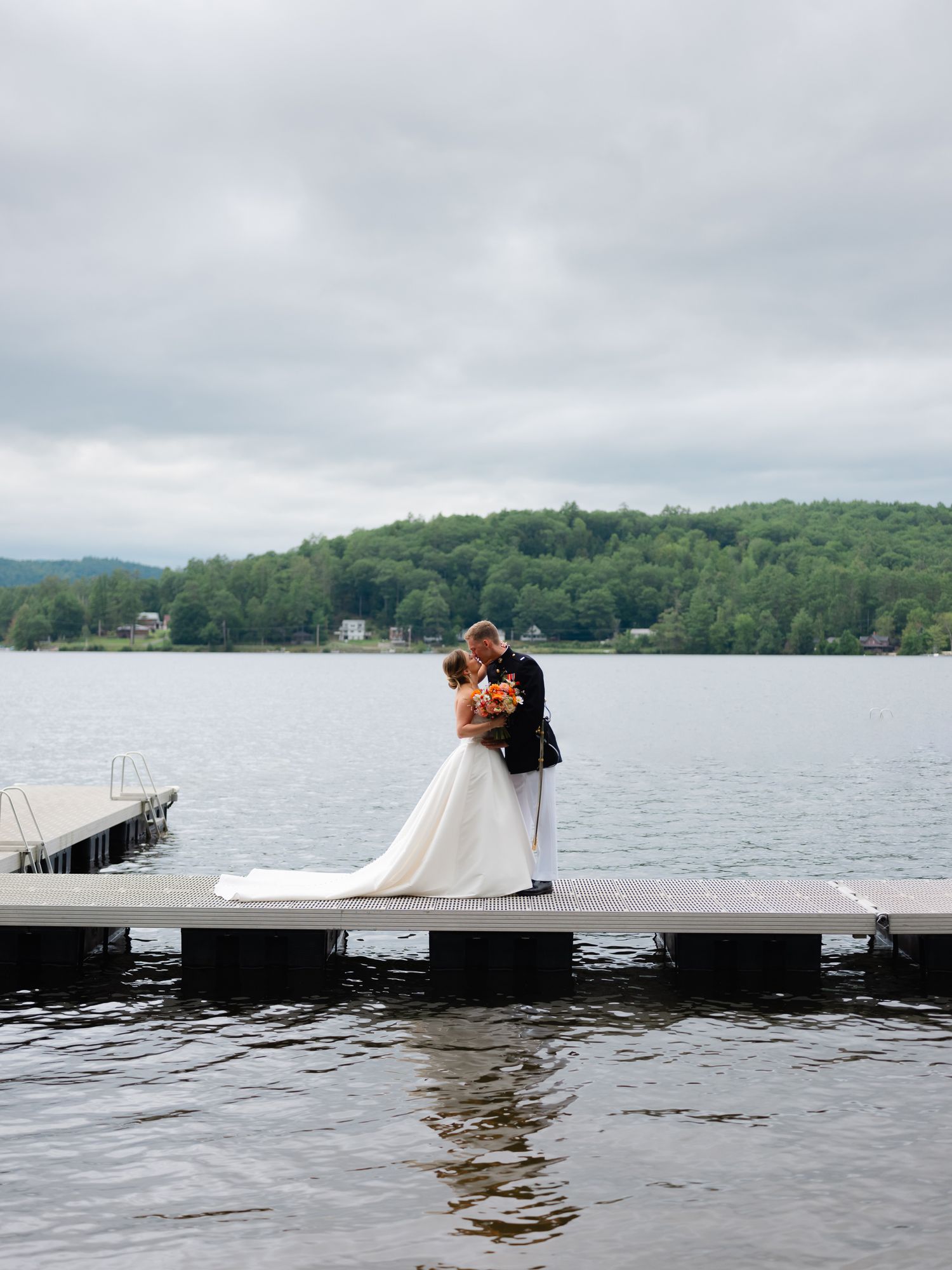 Bride and groom share romantic kiss on wooden dock overlooking calm lake with forested hills behind.