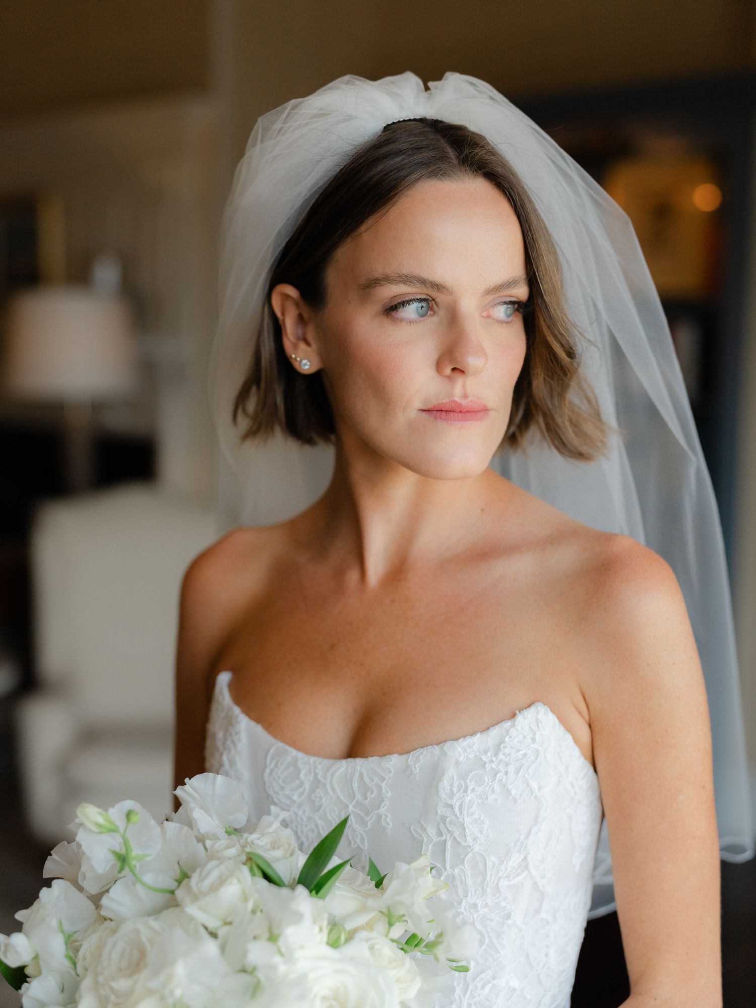 Bride with short brunette hair in strapless lace gown holds white bouquet gazing sideways.