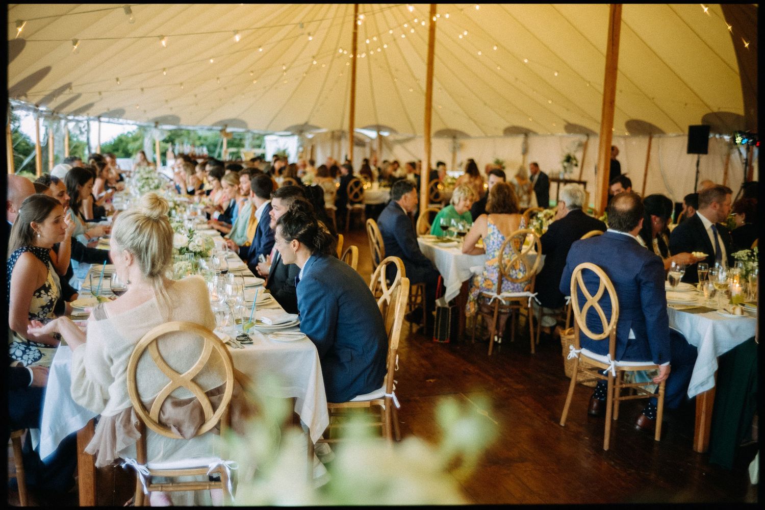 Wedding reception guests dine at long tables under a beautifully lit sailcloth tent.