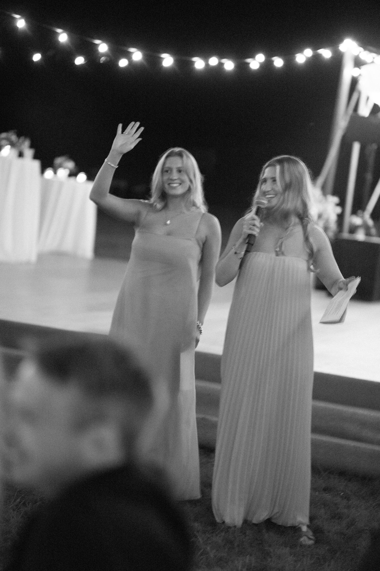 Two bridesmaids in elegant maxi dresses giving a speech with a microphone under string lights at an outdoor wedding reception.