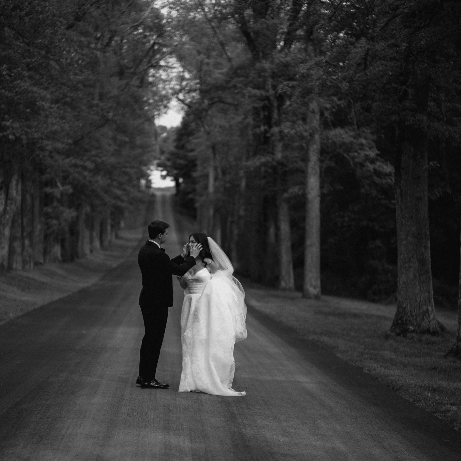 Black and white romantic photo of bride and groom facing each other on a tree-lined road.