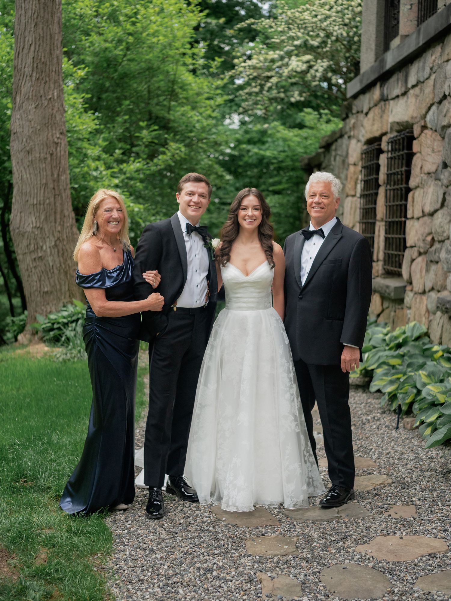 Newlyweds pose with family members in formal attire along a garden path at wedding venue.