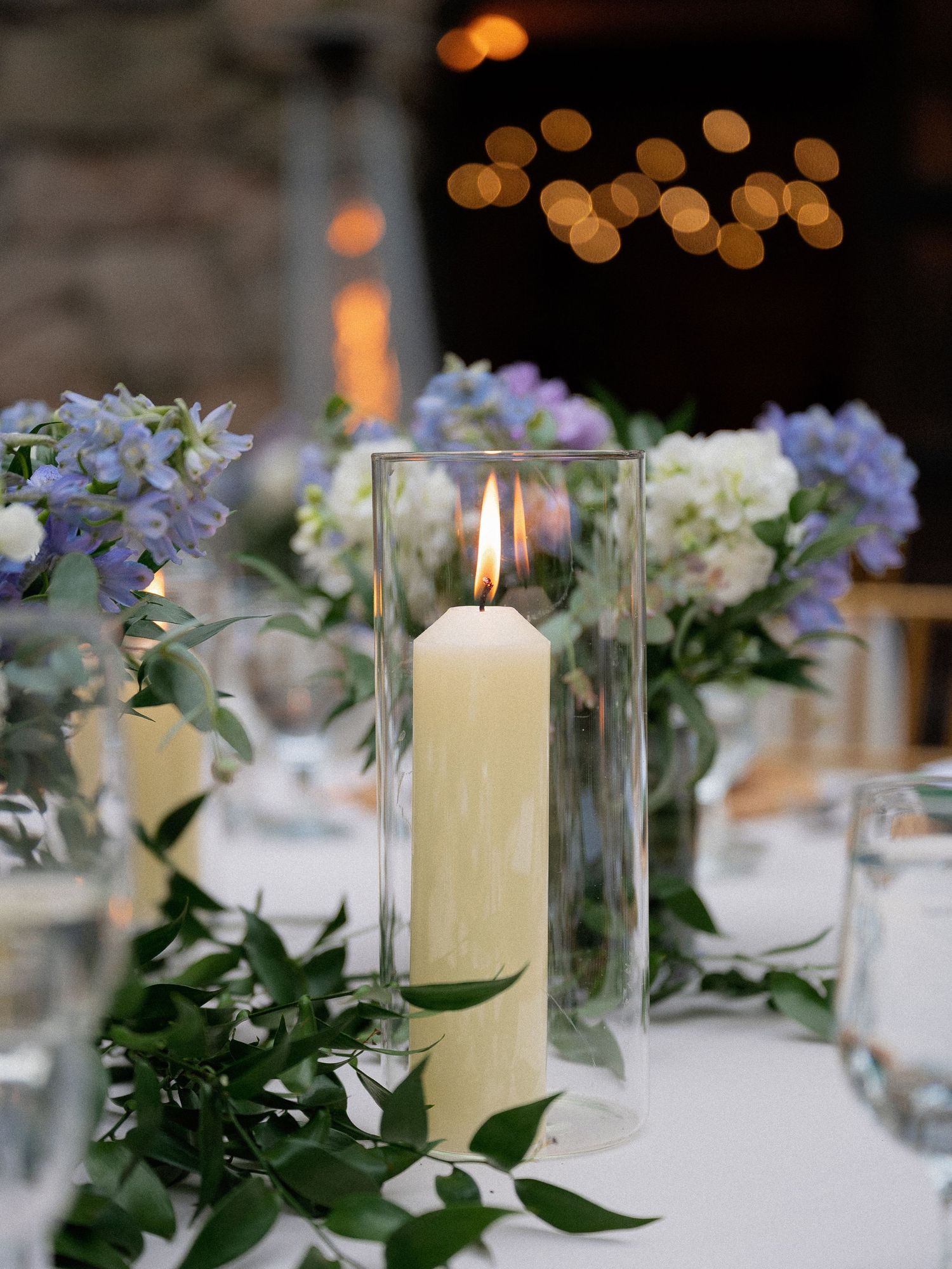 Lit pillar candle in glass hurricane vase surrounded by blue hydrangeas and greenery at wedding reception.