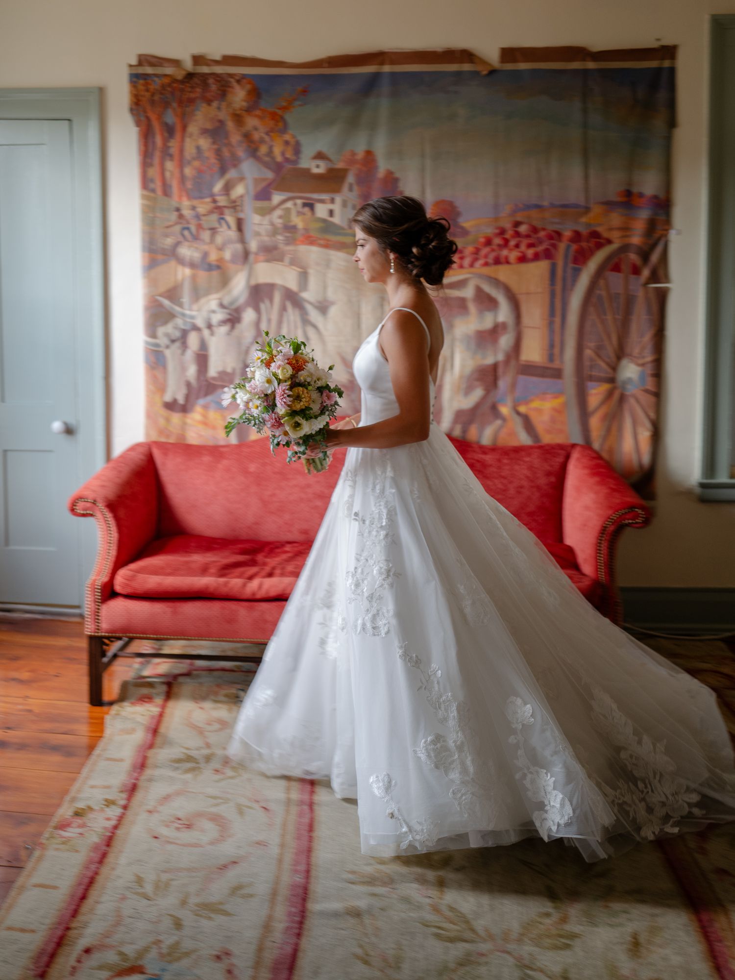 Bride in a lace ball gown holding a bouquet, standing in profile before a colorful folk art tapestry.