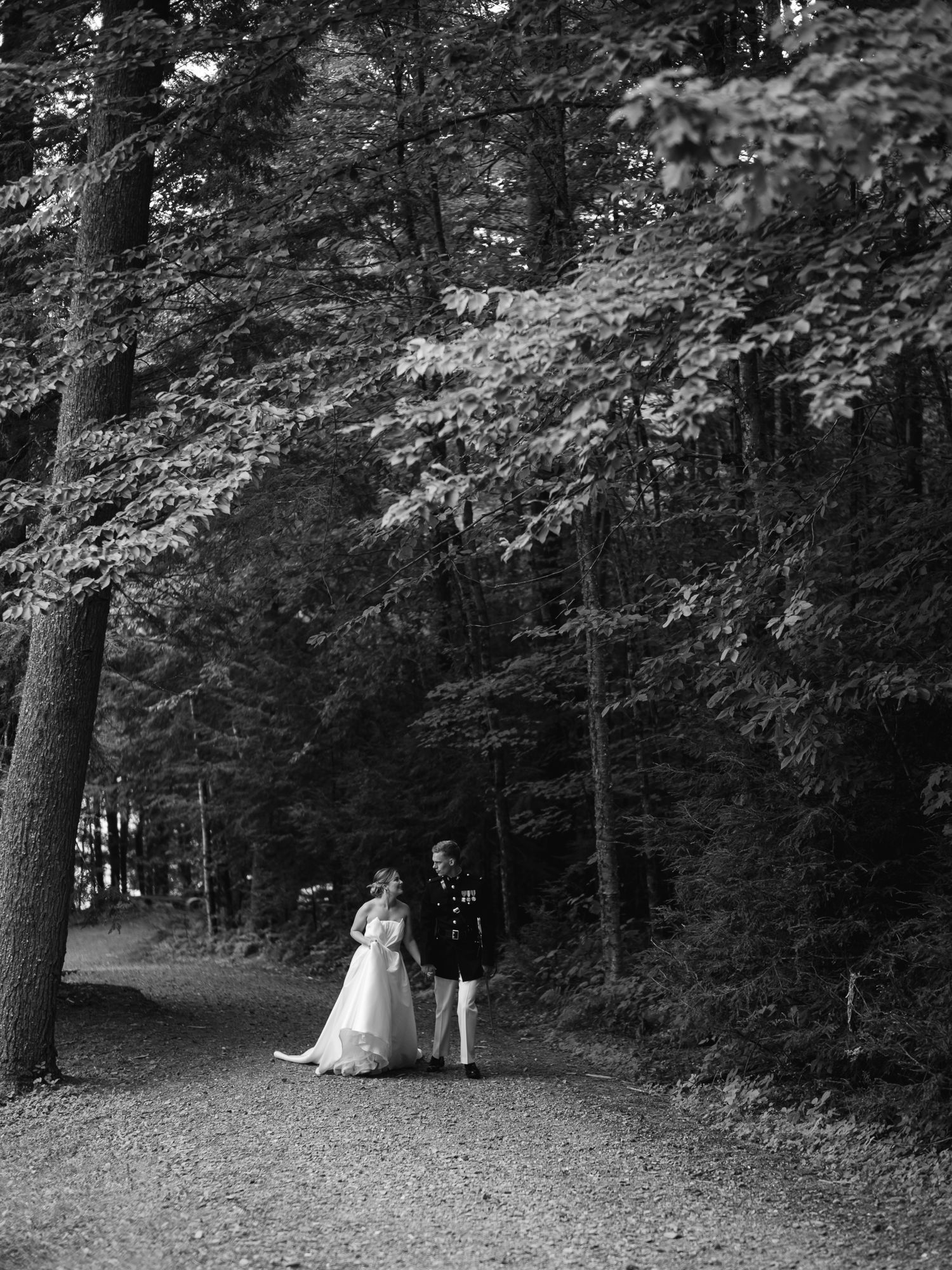 Black and white photo of bride and groom walking hand in hand down a tree-lined forest path.