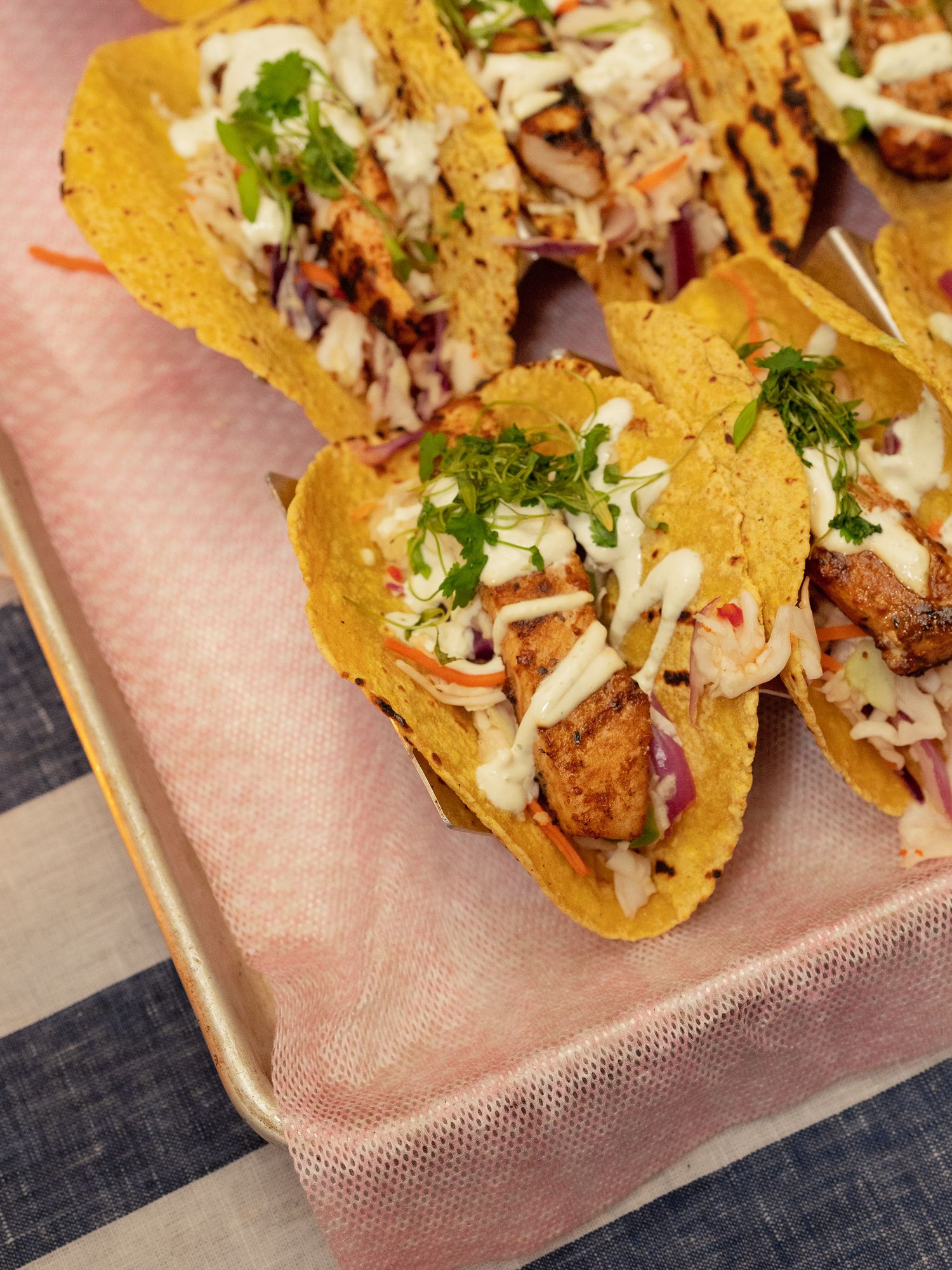 Crispy fish tacos in corn shells topped with coleslaw, cilantro, and creamy white sauce on a tray.
