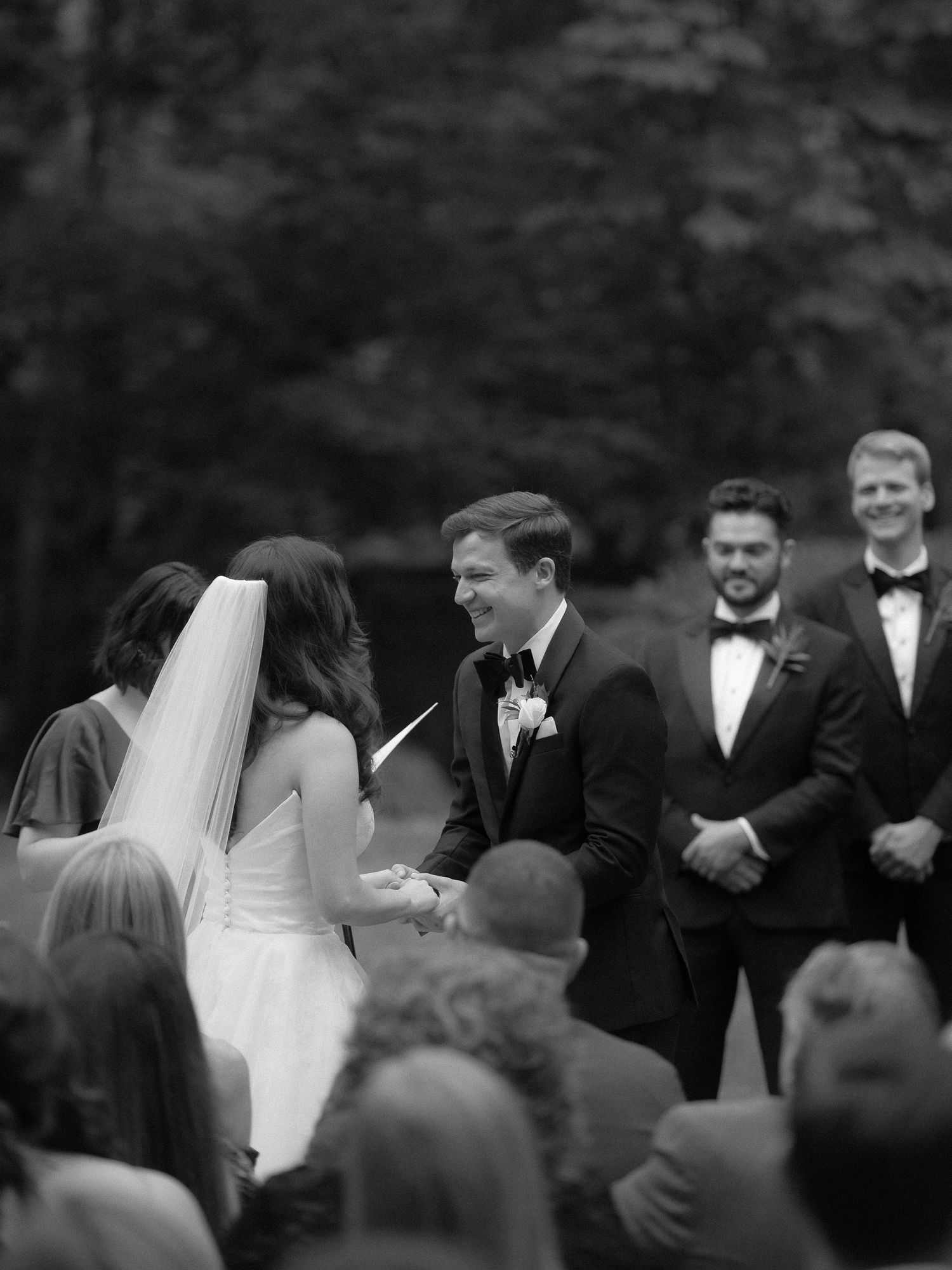 Black and white photo of a groom smiling at his bride during outdoor wedding ceremony vows.